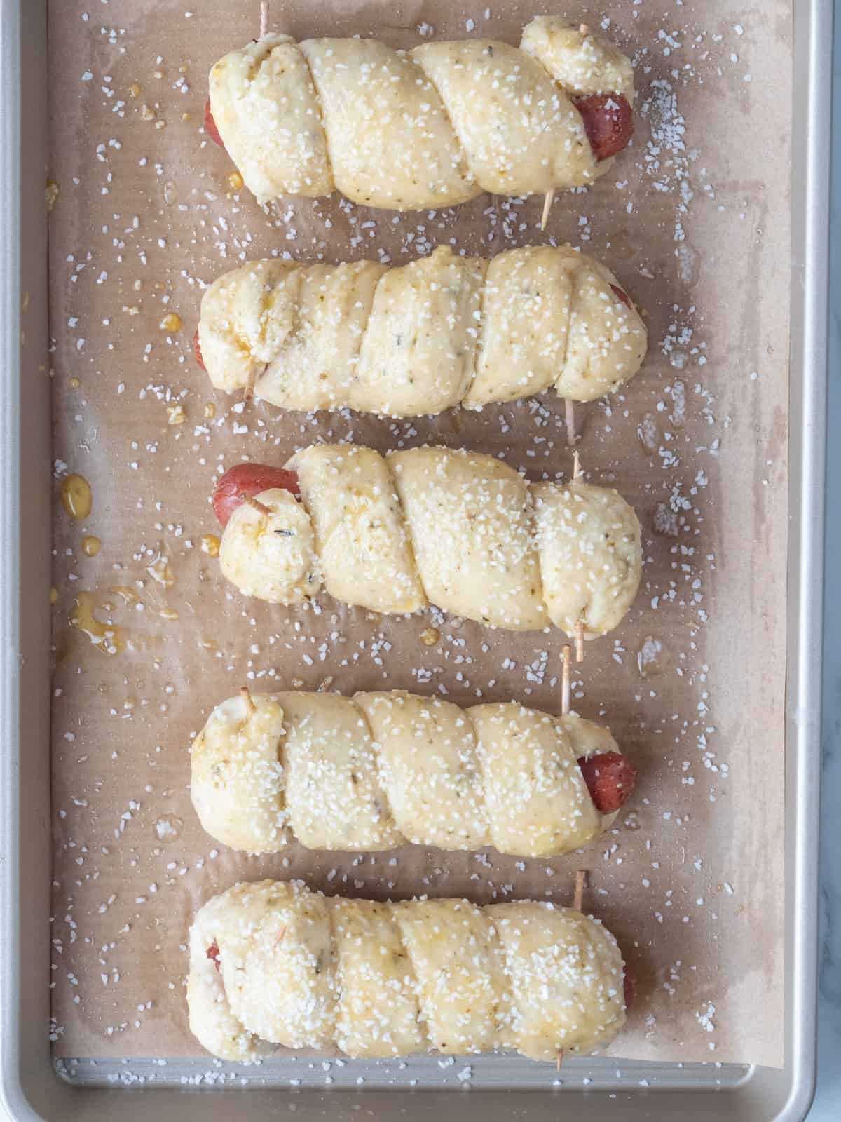 A parchment lined and greased baking sheet with boiled pretzel dogs brushed with a beaten egg yolk and water mixture and sprinkled with pretzel salt. 