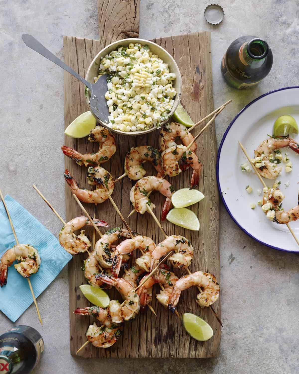 A wooden board with grilled mexican shrimp skewers with a bowl of corn salsa and lime wedges, along with a plate to serve and two beers next to it.