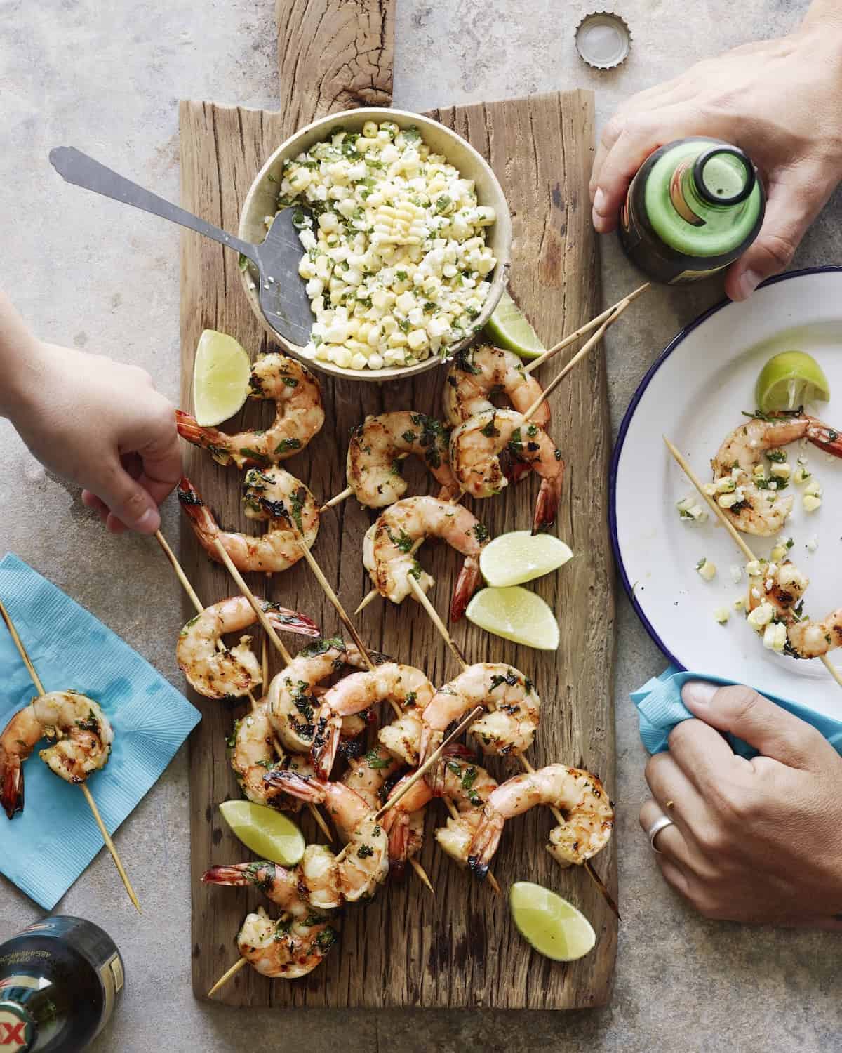 A wooden board with grilled mexican shrimp skewers with a bowl of corn salsa and lime wedges, along with a plate to serve and two beers next to it, along with a couple hands taking the skewers and beer.