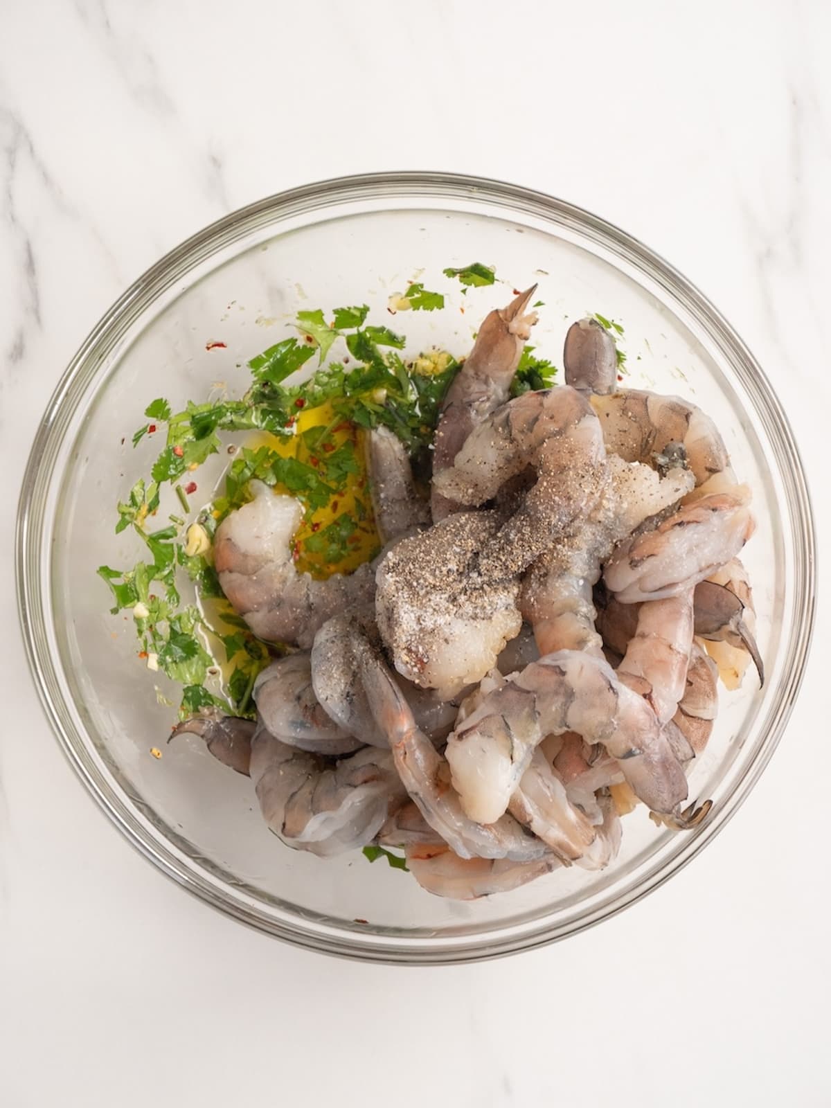 A small glass mixing bowl with an oil and herb mixture, and raw deveined peeled shrimp tossed in it and coated.
