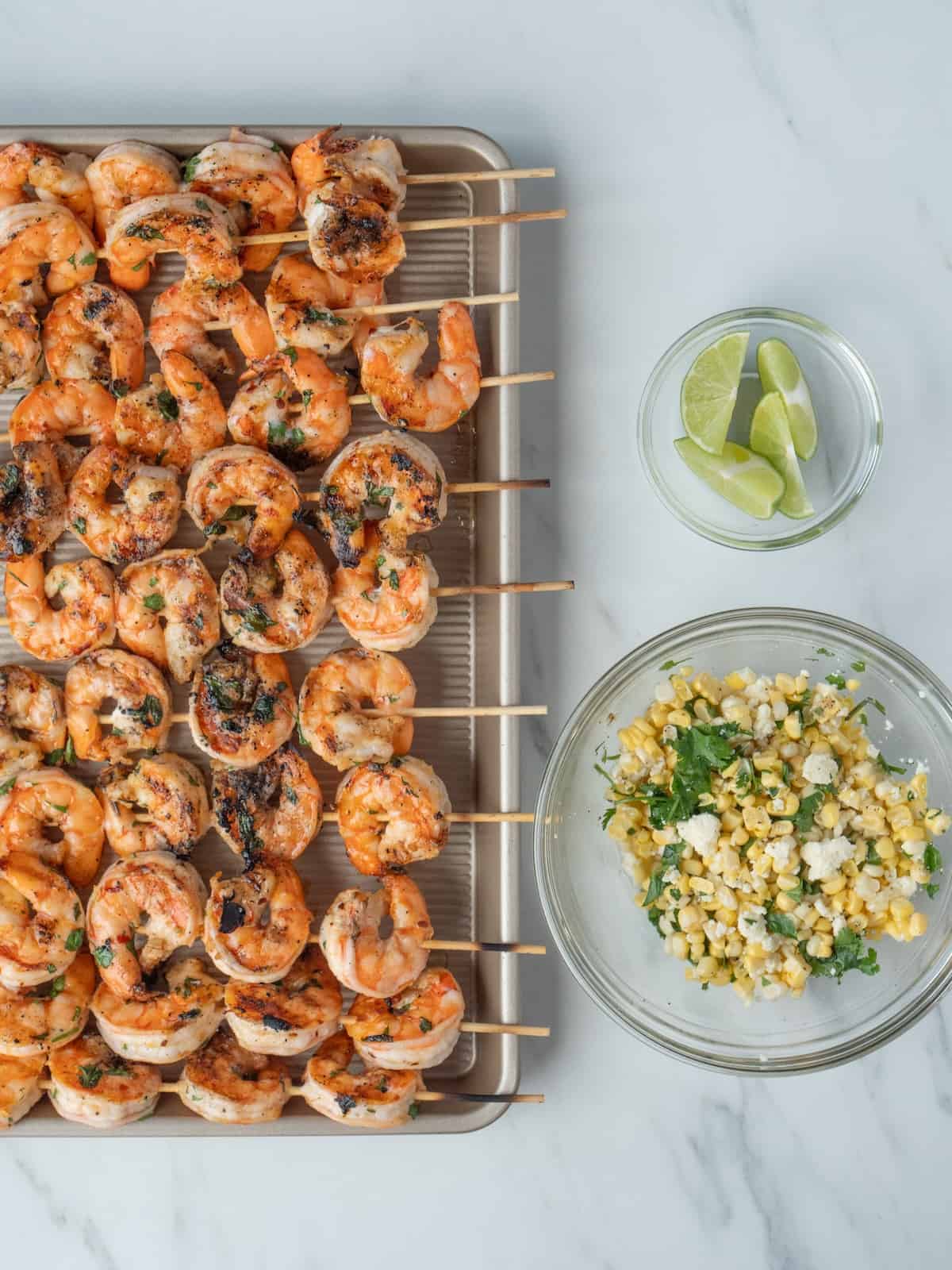 A baking sheet with grilled shrimp skewers, along with a glass bowl of corn salsa, and another one with lime wedges.