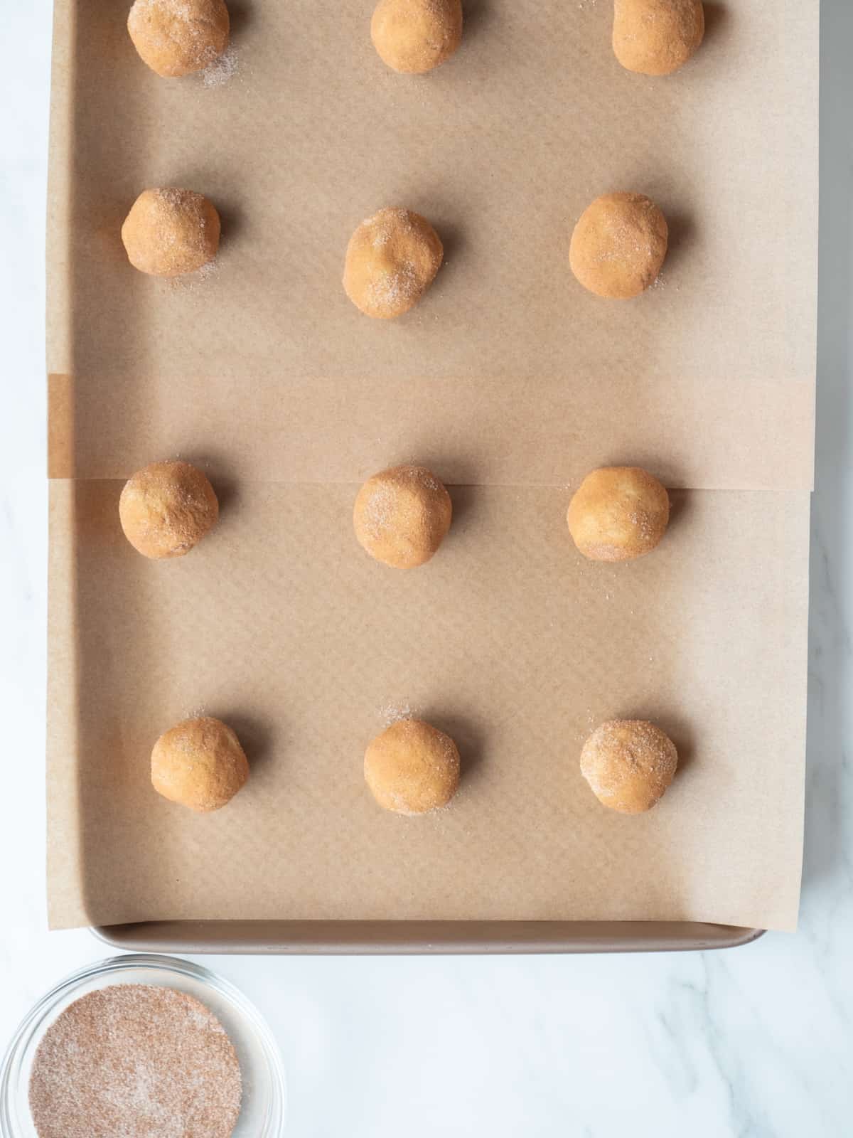 A parchment paper lined baking sheet with twelve snickerdoodle cookie dough balls, rolled in a mix of cinnamon and sugar and placed spaced from each other in a grid, with a glass bowl of cinnamon and sugar mixed together on the side.