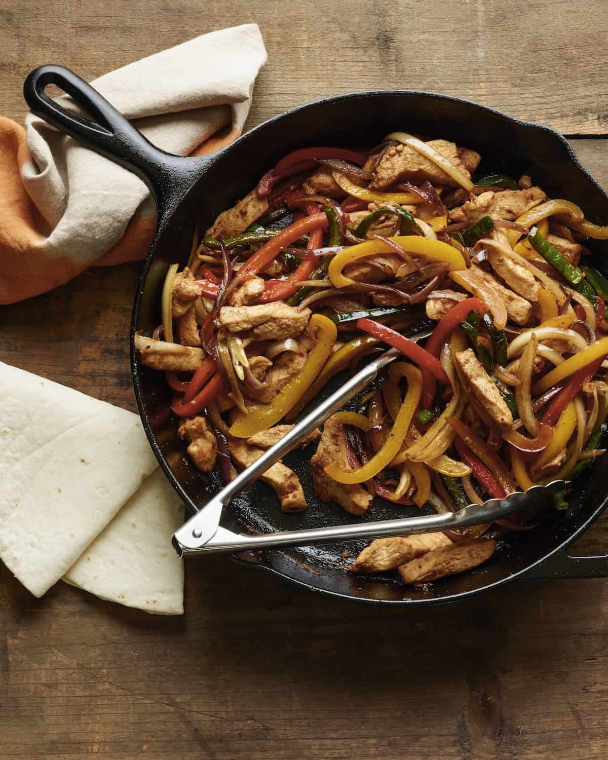A cast iron with poblano pepper chicken fajitas with tongs to serve, along with a folded tortillas and a kitchen towel to hold the cast iron.