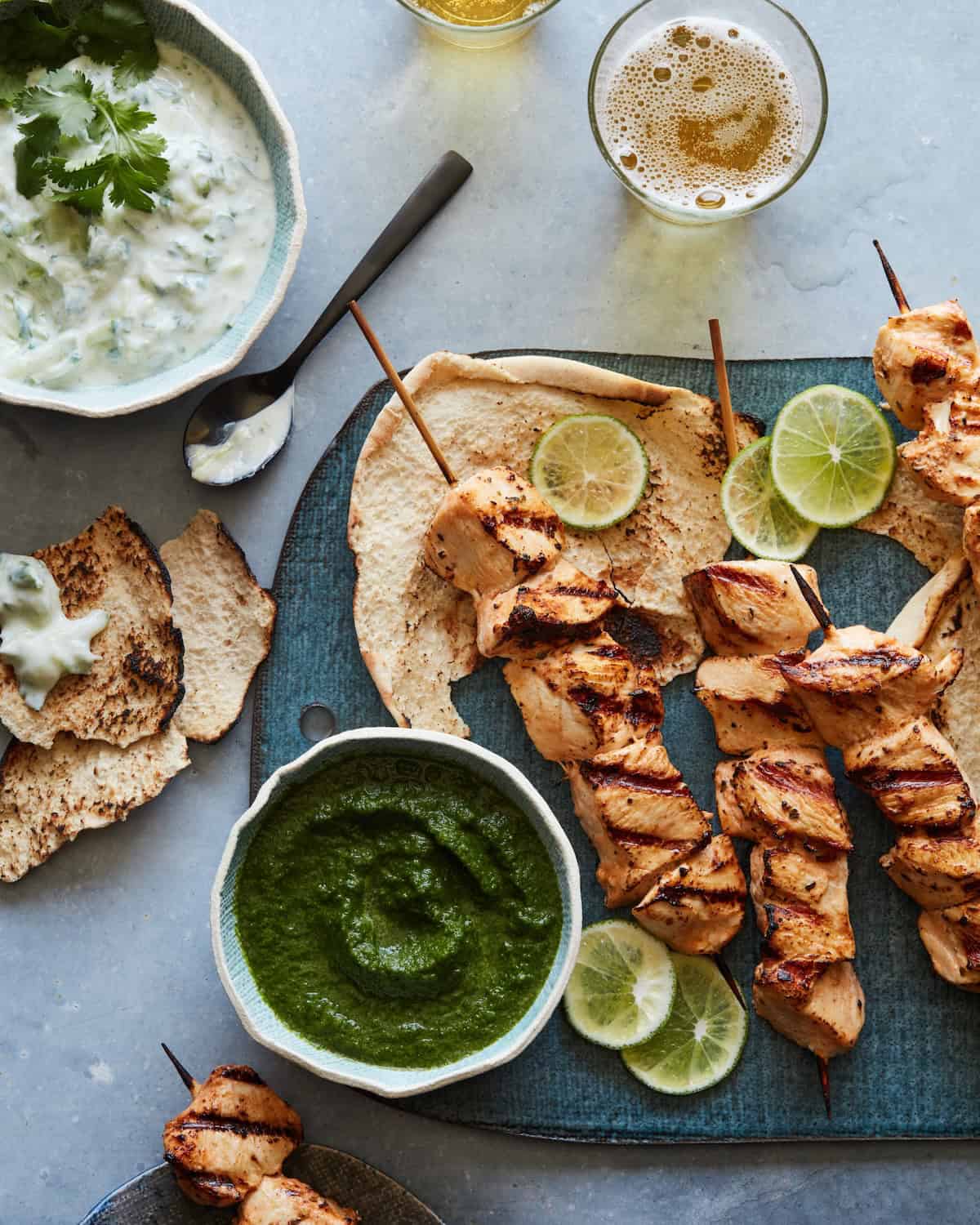 Chicken skewers on a board topped with thinly sliced limes and some pita halves, with a small bowl of cilantro mint sauce and a bowl of tzatziki on the side.