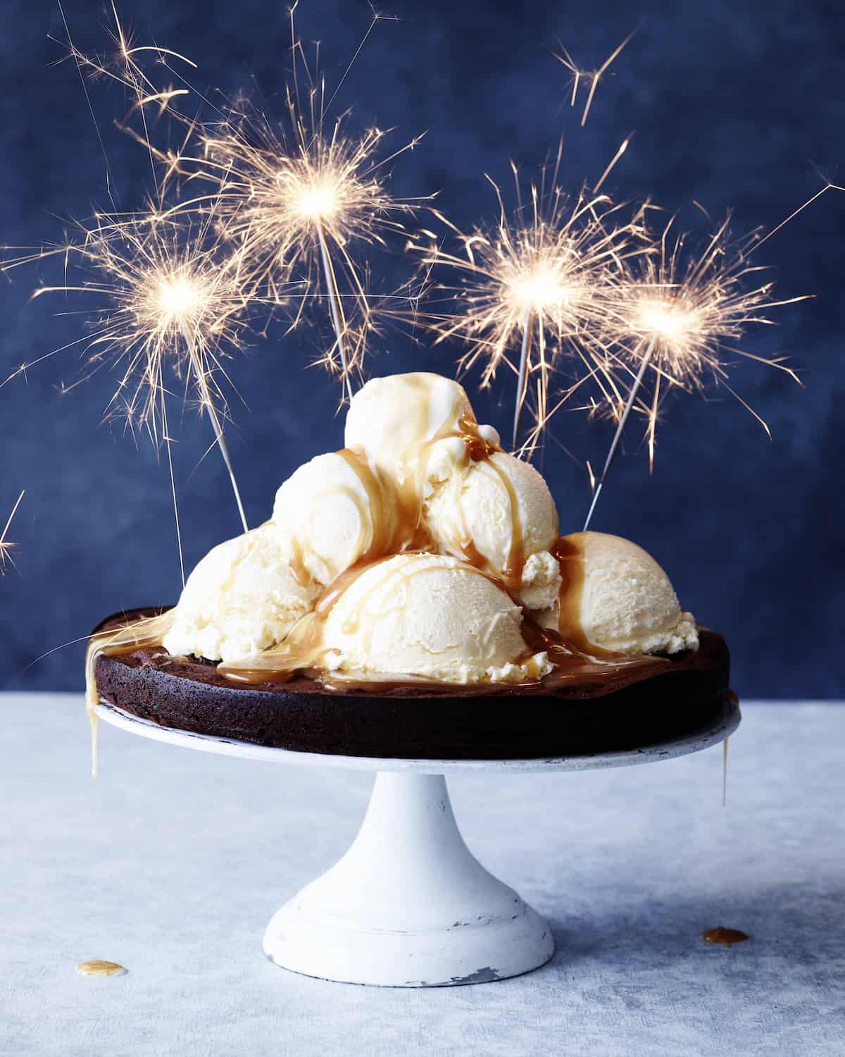 A flourless chocolate cake on a white cake stand, topped with caramel sauced and heaps of vanilla ice cream scoops, along with sparkler candles that are lit up.