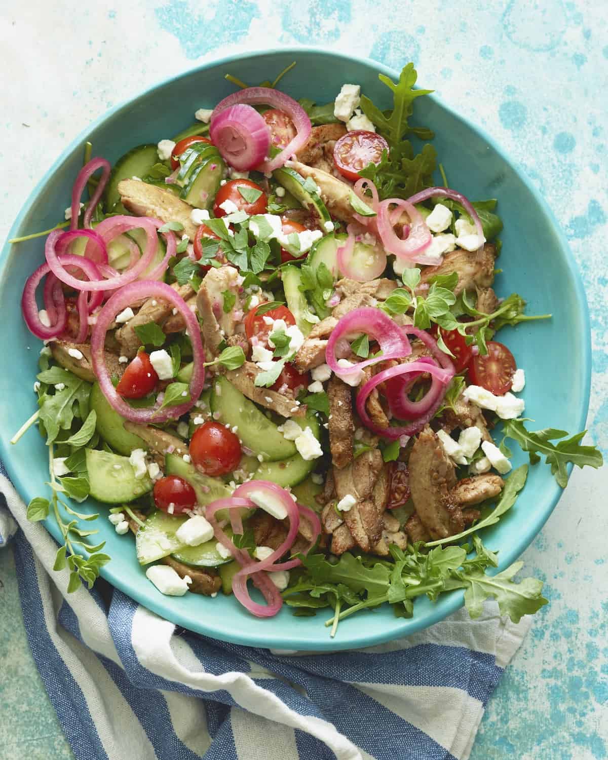 A shallow bowl with a greek chicken salad, topped with crumbled feta and pickled onions, placed on a kitchen towel.