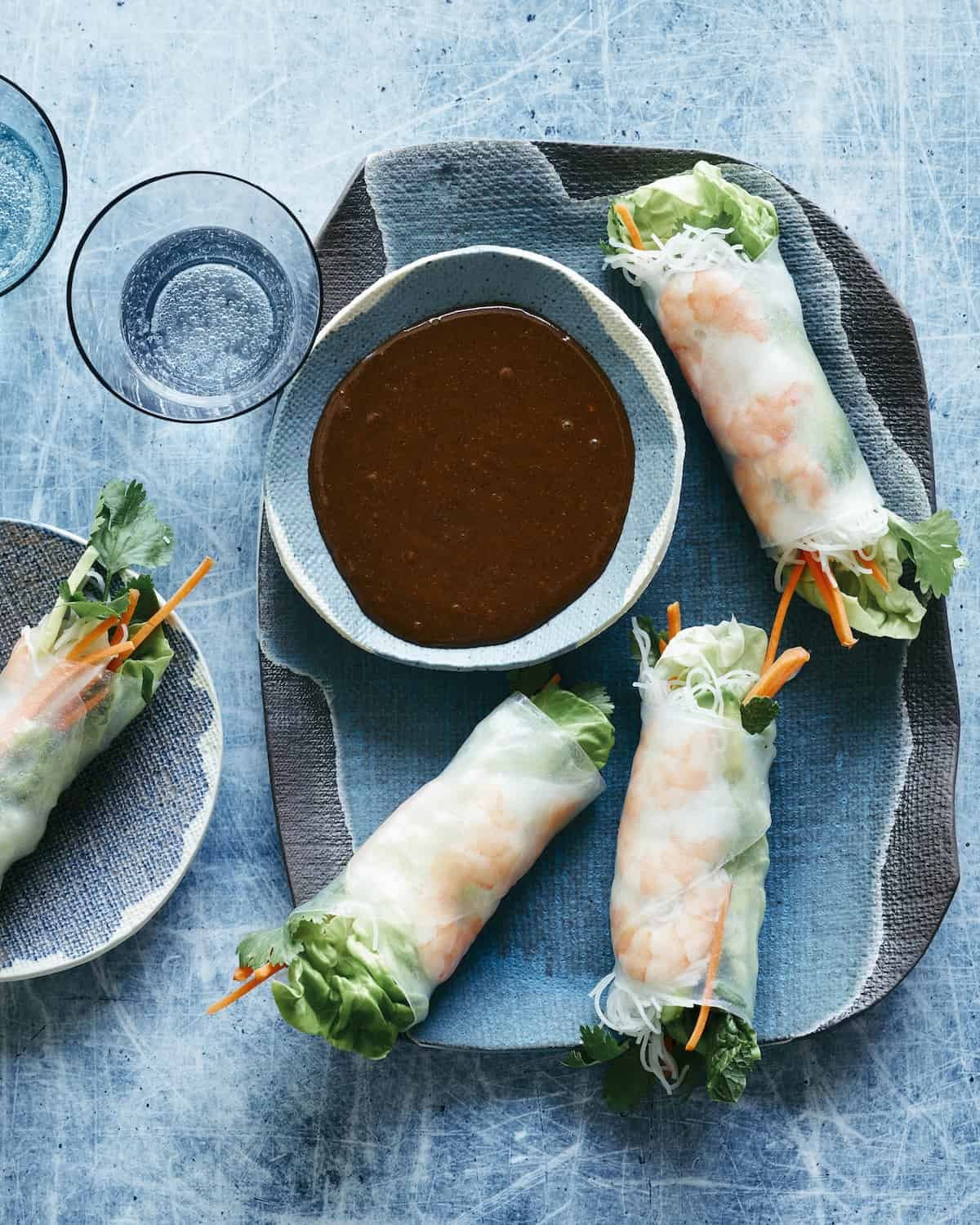 A rectangular platter with avocado shrimp spring rolls with a bowl of hoisin-peanut butter sauce for dipping, and another small plate with a spring roll served in it.