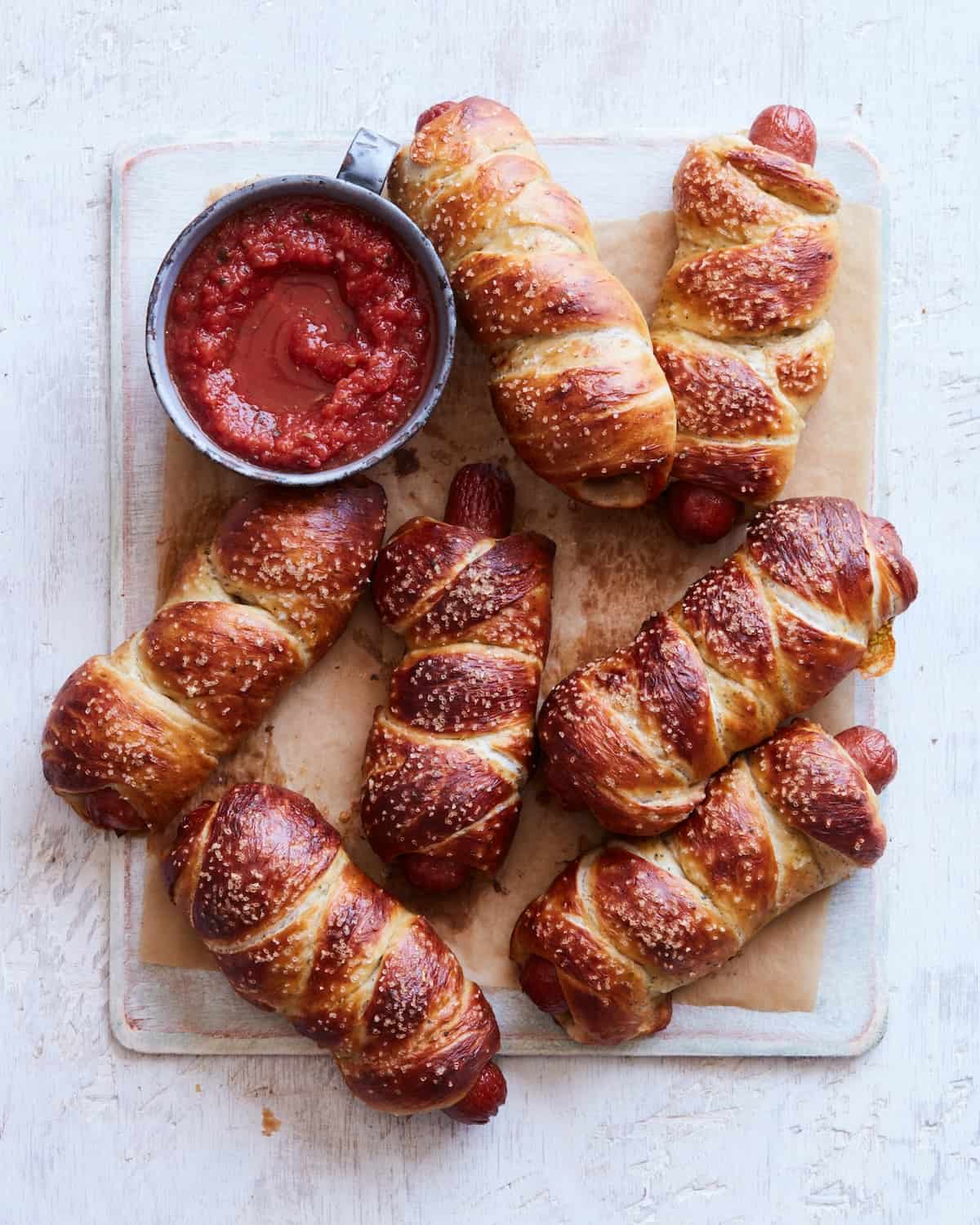 Seven pretzel dogs and a small bowl of marinara sauce for dipping placed on a parchment paper lined board.