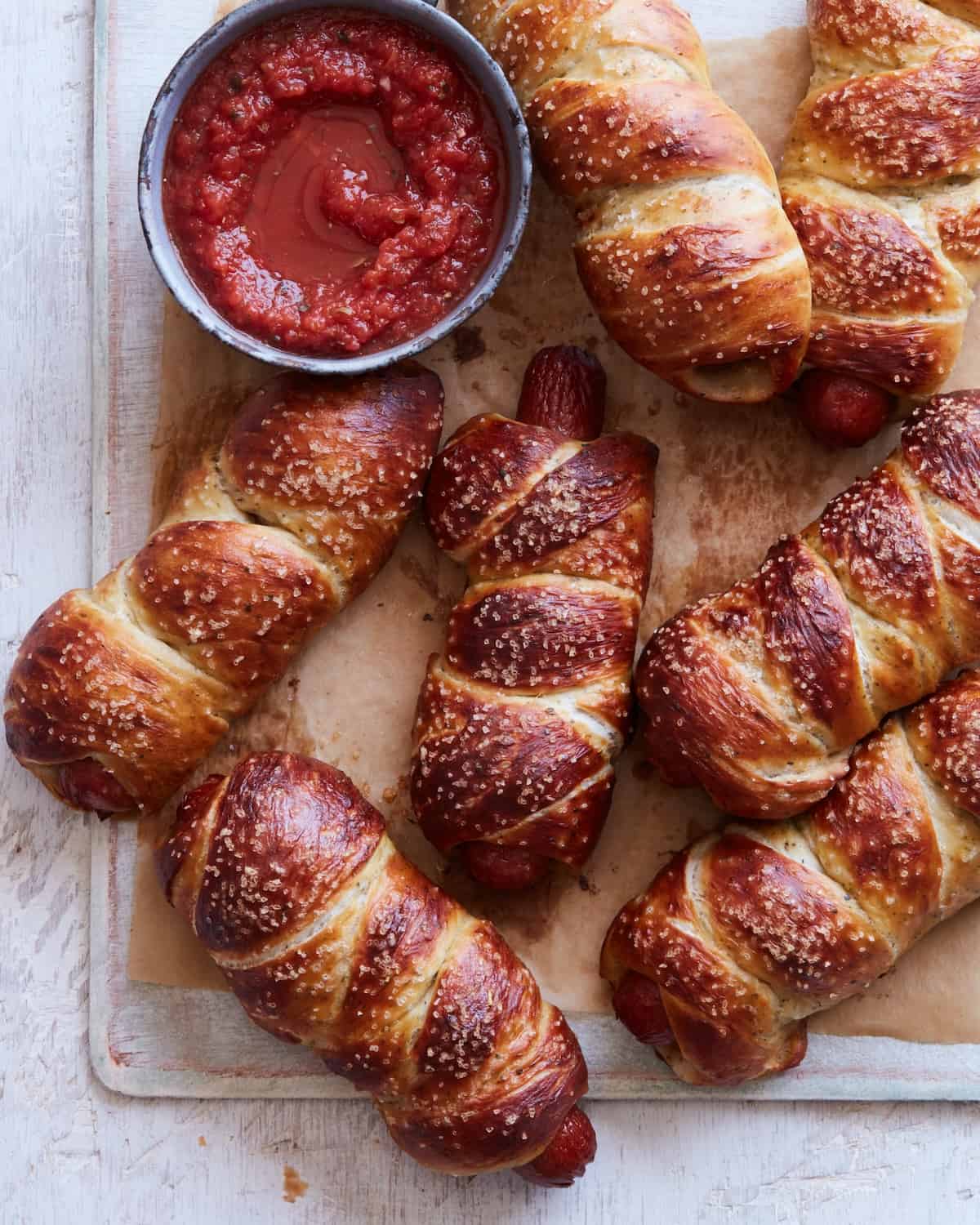Seven pretzel dogs and a small bowl of marinara sauce for dipping placed on a parchment paper lined board.