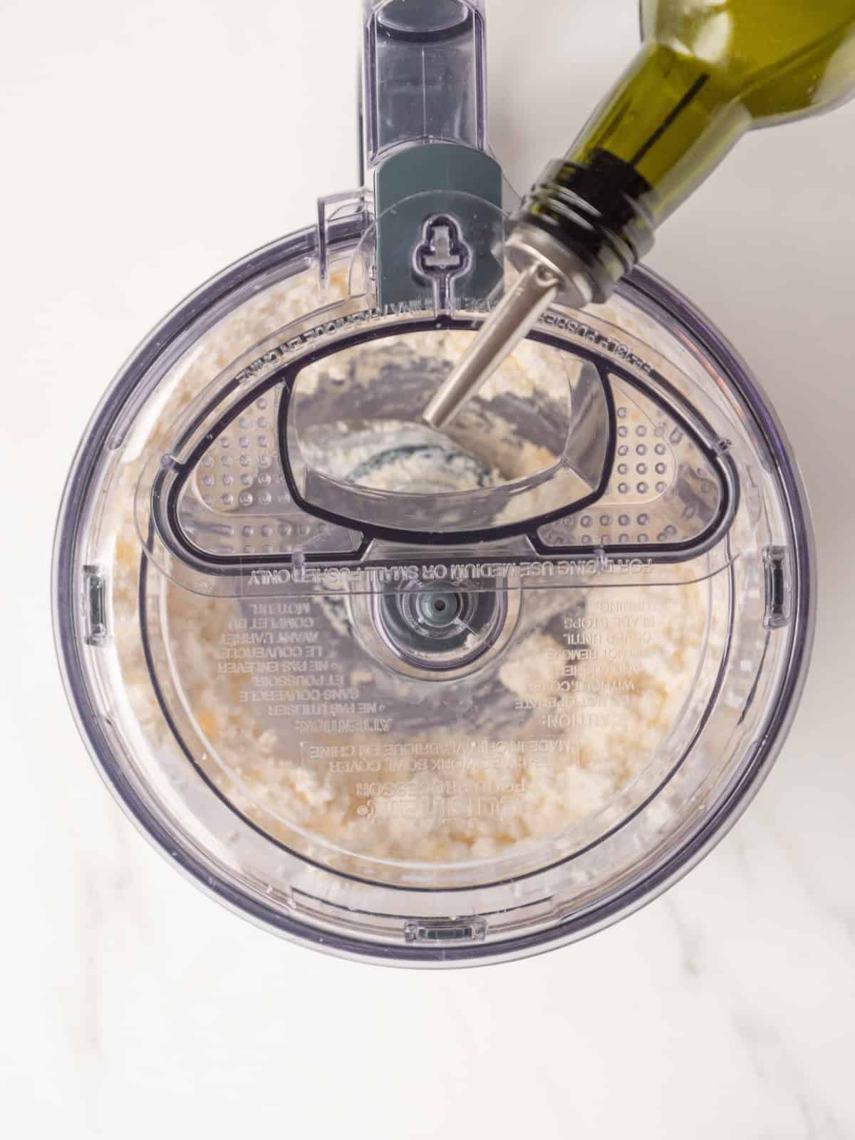 A blender with whipped feta being made, and extra virgin olive oil being drizzled through the opening on top.
