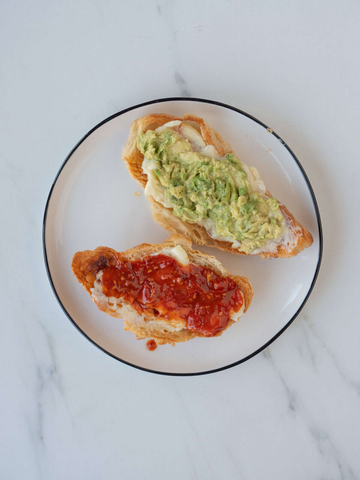 A plate with two toasted halves of a croissant, one with mayonnaise and tomato jam spread, and the other half spread with mayonnaise and smashed avocado.