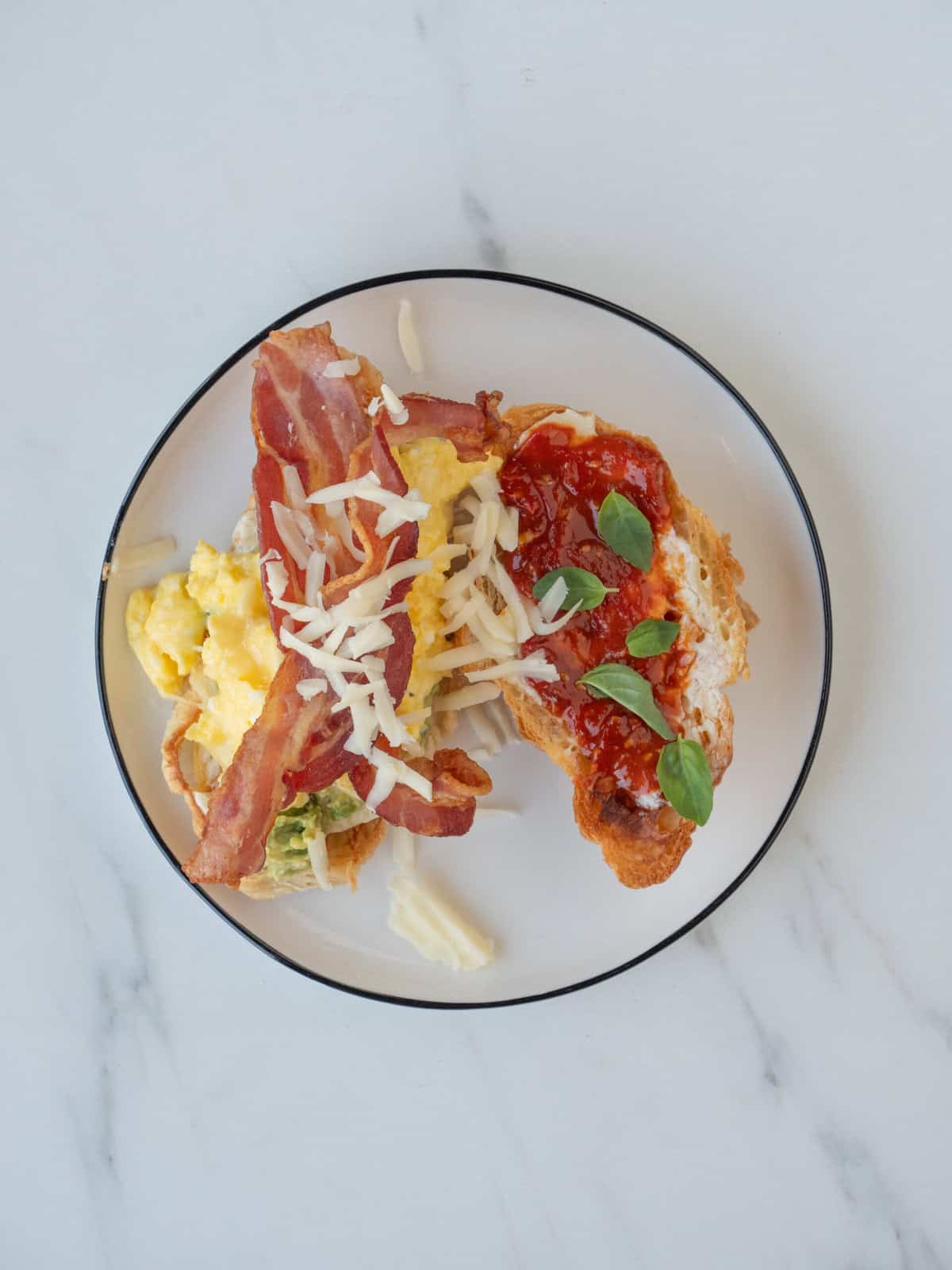 A plate with two toasted halves of a croissant, one with mayonnaise, tomato jam and basil leaves, and the other half spread with mayonnaise, smashed avocado, soft scrambled eggs, bacon and shredded fontina.