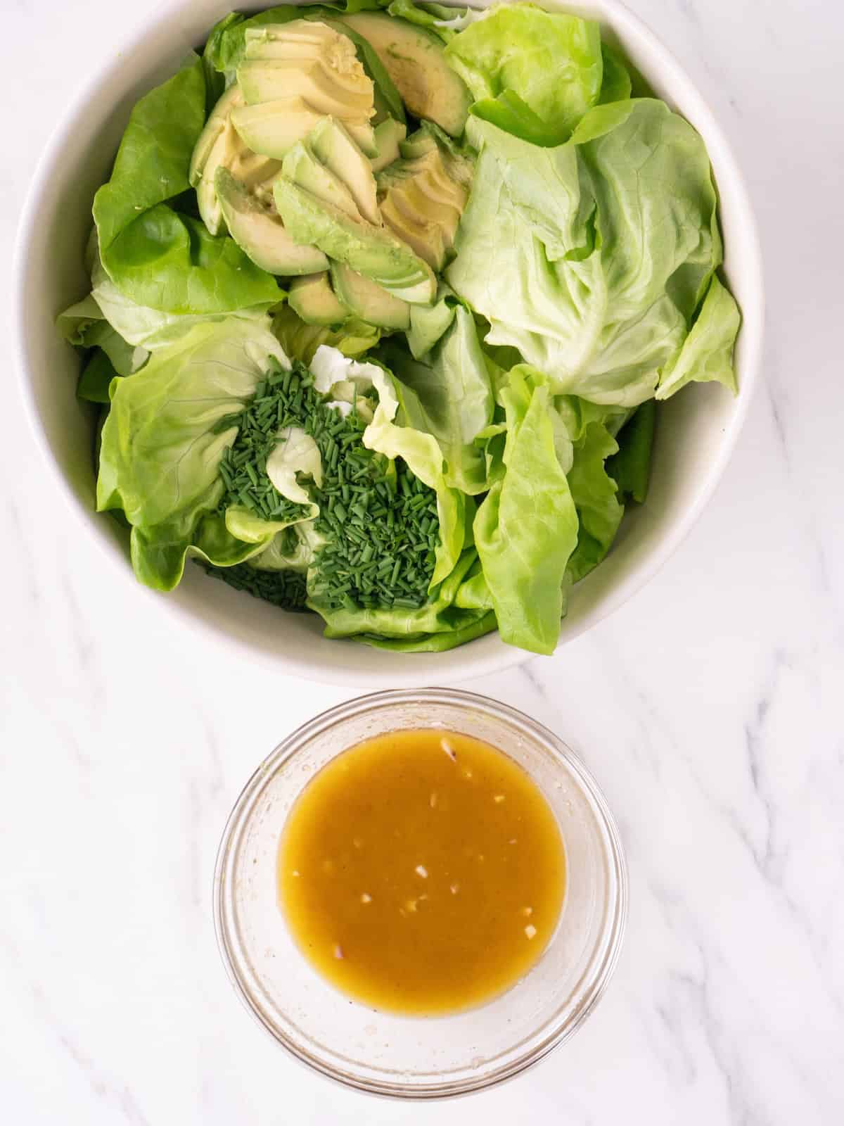 A bowl with butter lettuce, chopped chives and sliced avocado to make a butter lettuce salad, along with a glass bowl with lemon shallot vinaigrette on the side.