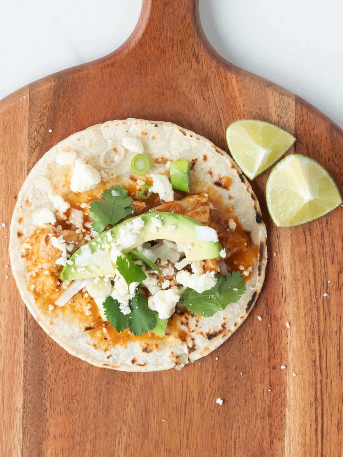 A wooden board with a lightly charred tortilla with chicken tinga topped with cilantro, chopped onions, sliced scalions, avocado and crumbled cotija cheese added along with lime wedges on the side.