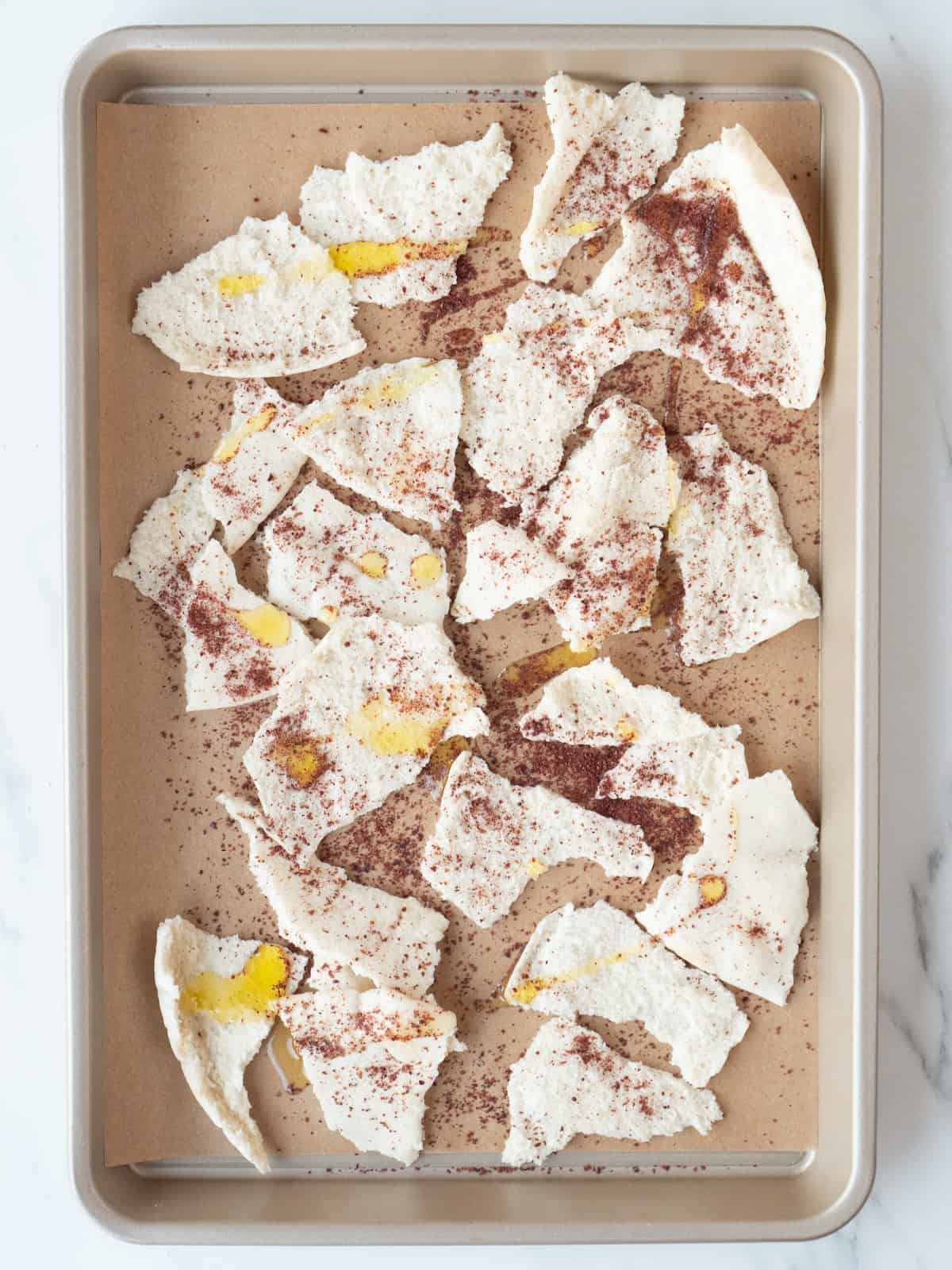 A parchment paper lined baking sheet with pita torn into pieces, drizzled with olive oil and sprinkled with sumac.