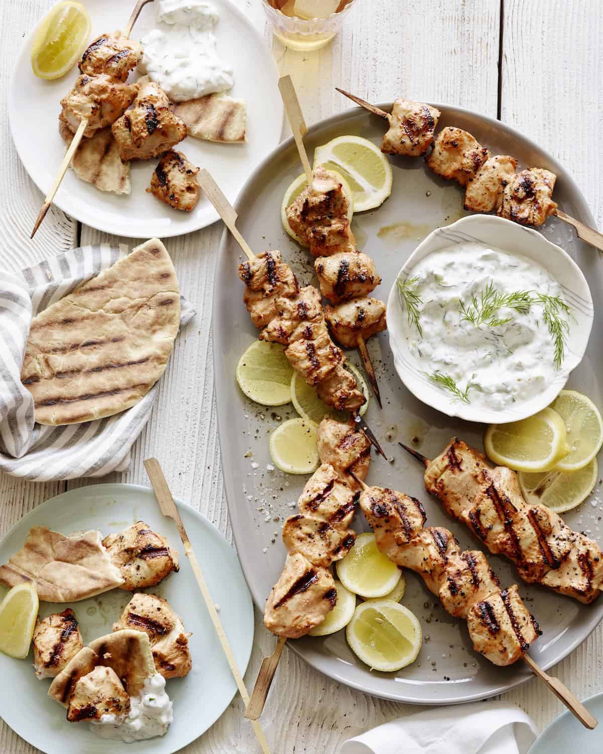 A platter with yogurt marinated chicken skewers along with lemon slices, a bowl with tzatziki and plates on the side with the skewers served with pita.