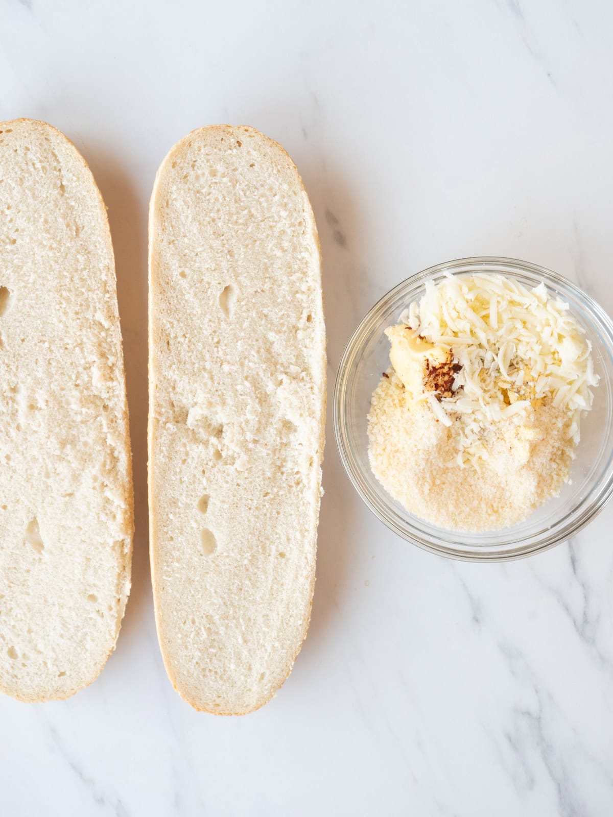An italian or french bread loaf cut into two halves horizontally, along with a small glass mixing bowl of butter, chopped garlic, red pepper flakes, shredded mozzarella, parmesan, salt and pepper.
