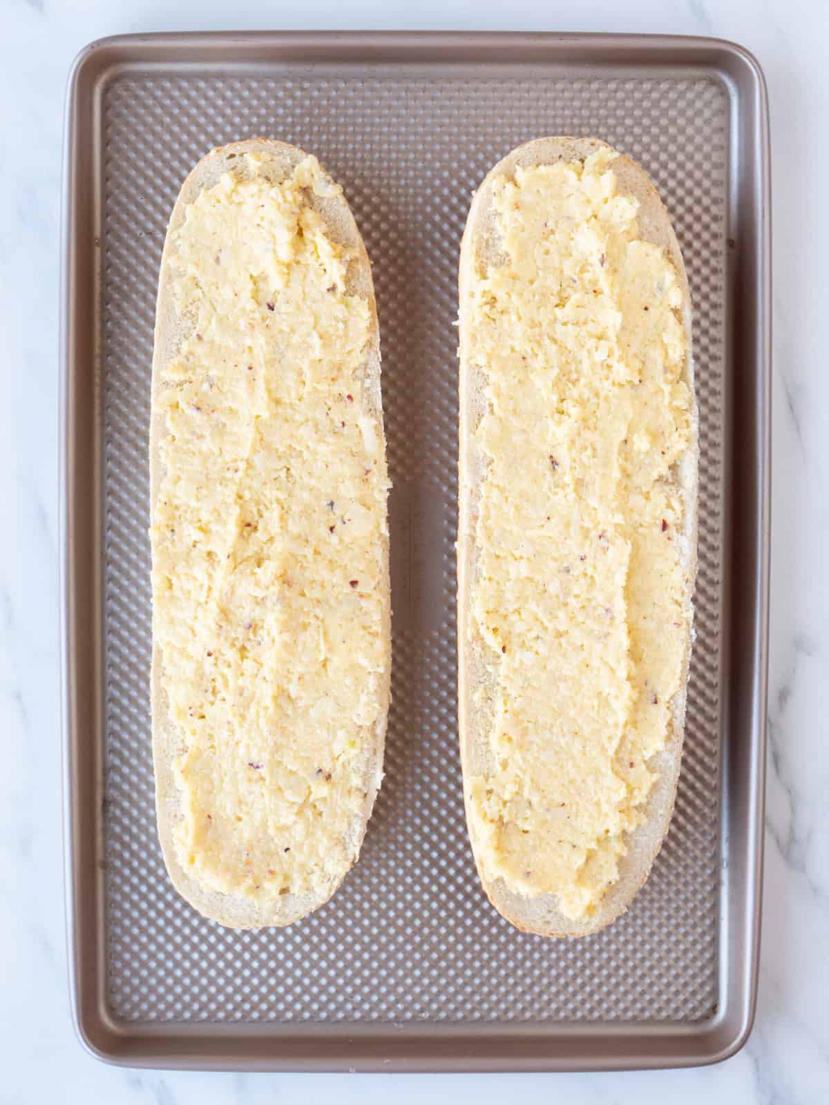 Horizontally cut halves of a bread loaf on a baking sheet, with the butter cheese garlic red pepper flakes mixture spread evenly on both, ready to bake.