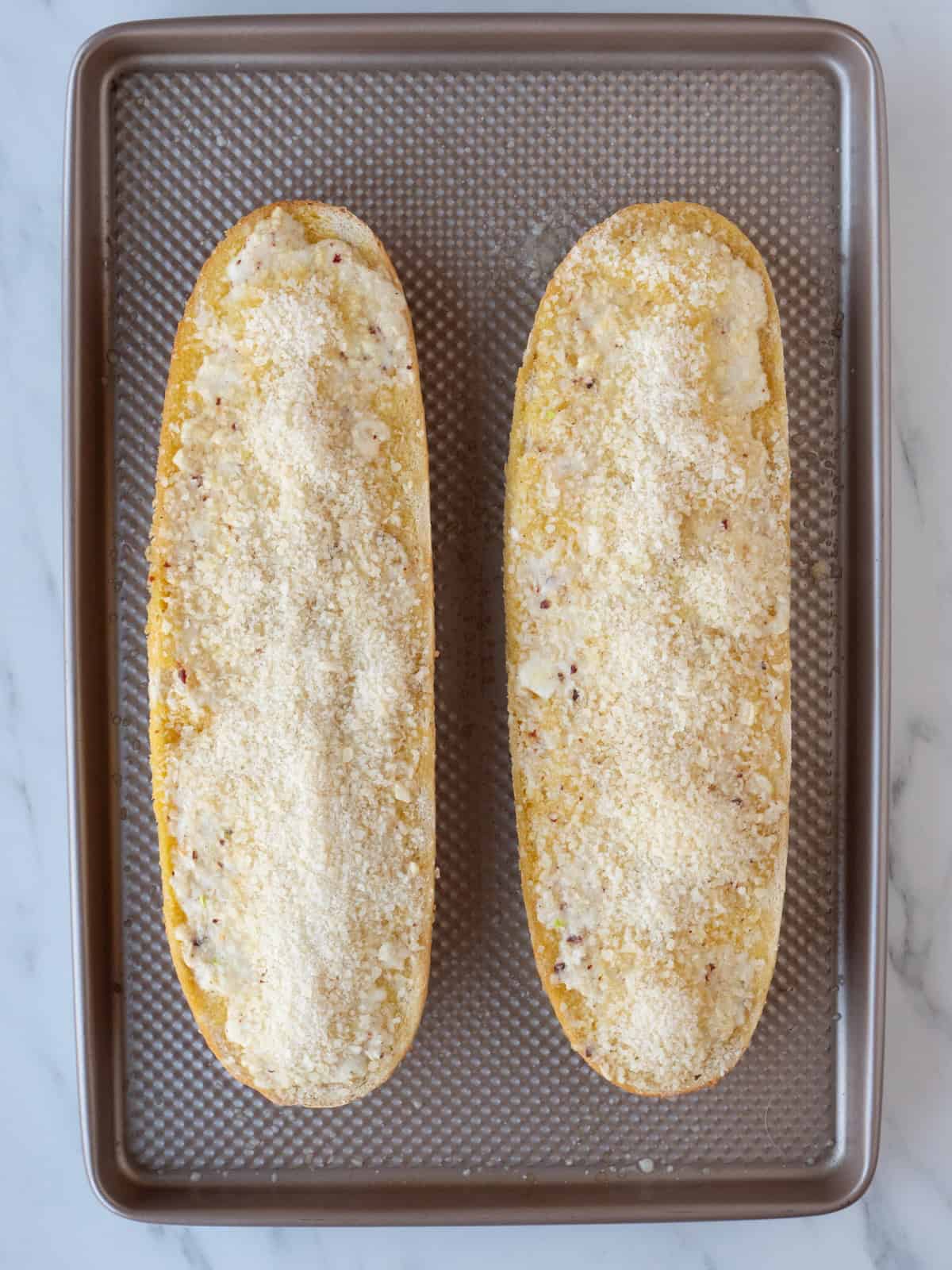 Horizontally cut halves of a bread loaf on a baking sheet, with the butter cheese garlic red pepper flakes mixture on them starting to melt, and topped with more parmesan ready to be put back in the oven.