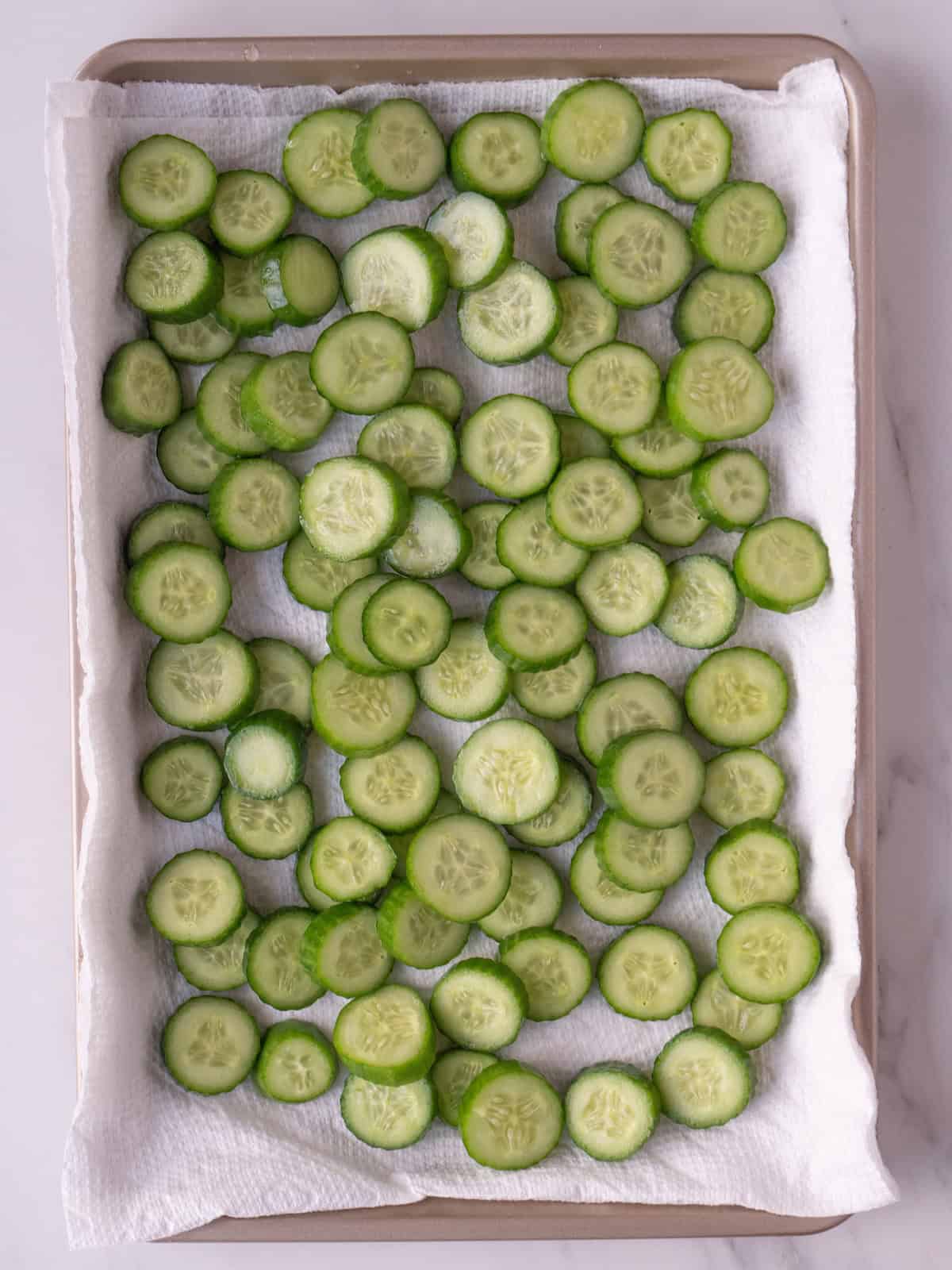 A baking sheet lined with paper towels with rinsed sliced cucumbers drying on it.