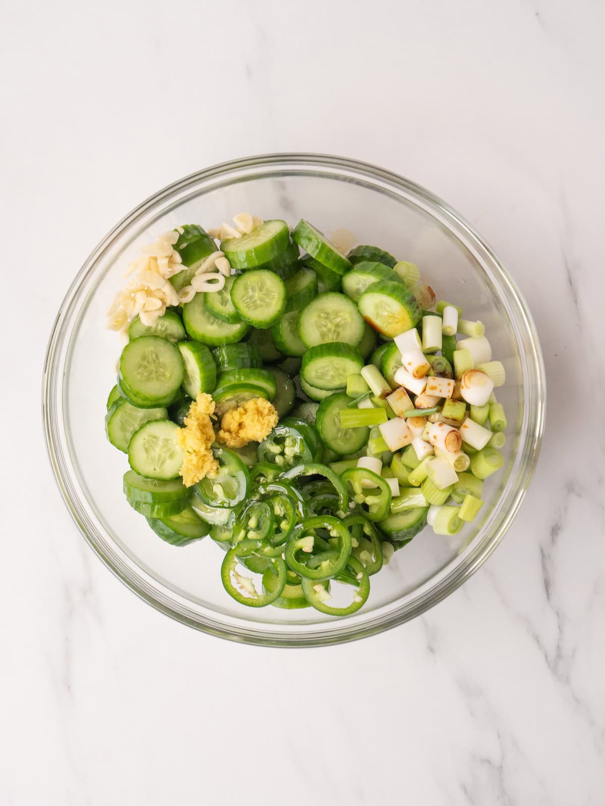 A glass bowl with sliced cucumbers, chopped green onions, jalapeños, ginger and garlic.