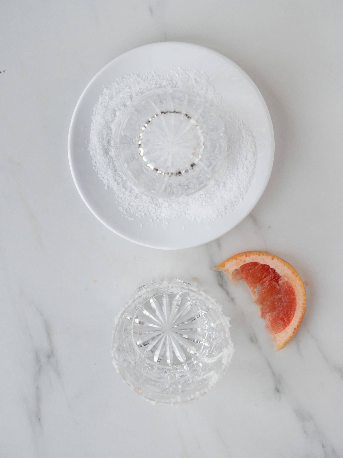 A small plate with salt, and a cocktail glass rim being dipped in it to salt its rim, with a grapefruit wedge and another salted rim cocktail glass on the side.