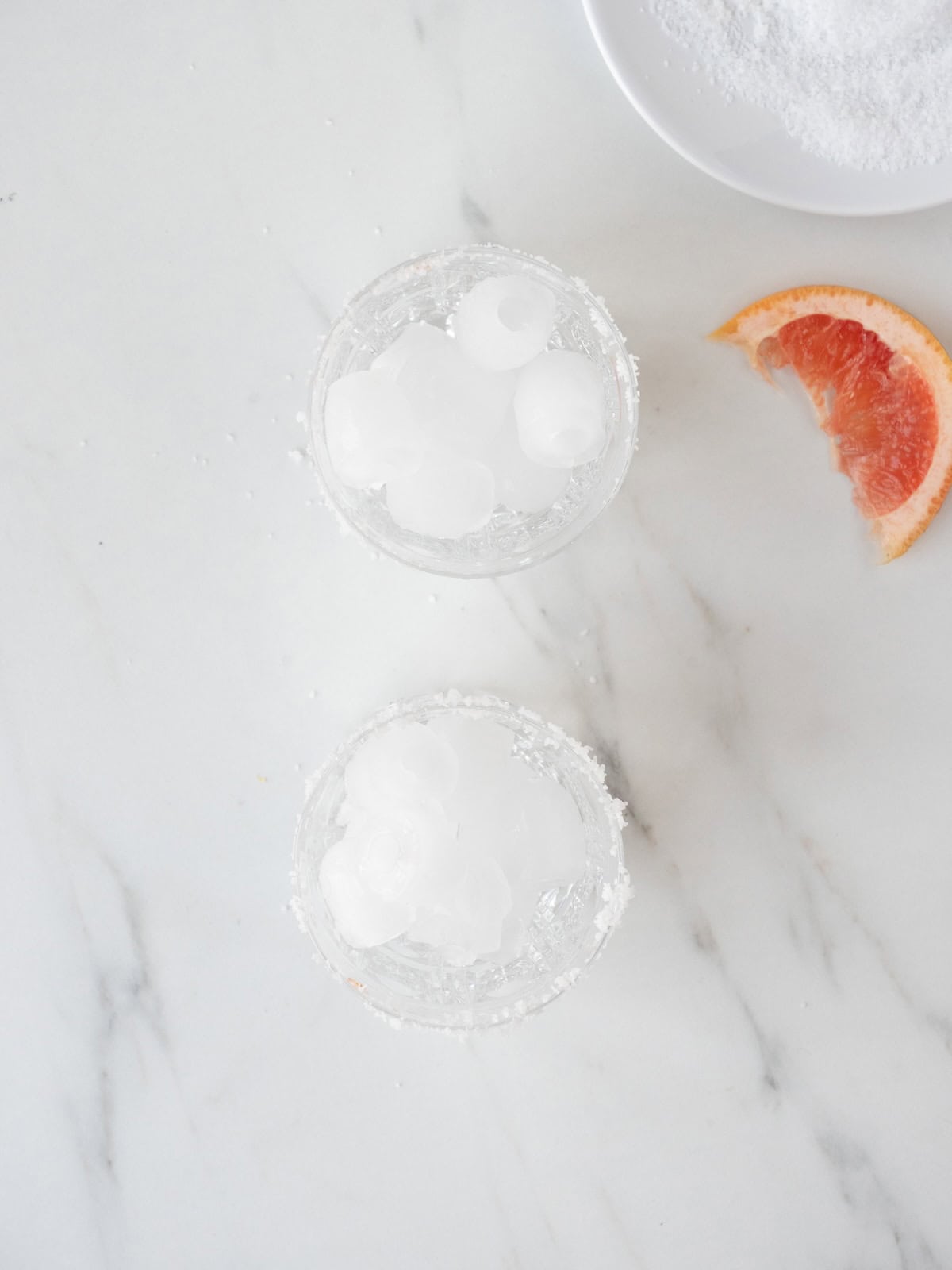 Two salt rim cocktail glasses with ice in them along with a grapefruit wedge and a small plate with salt on the side.