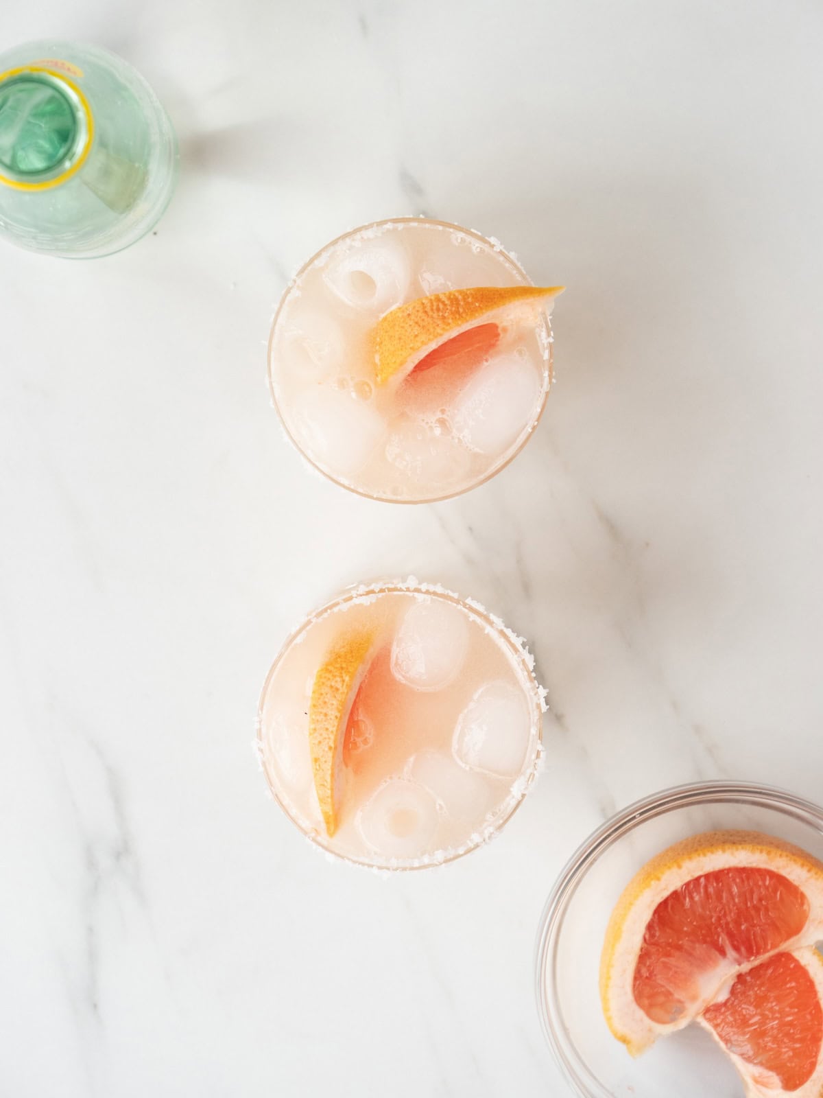 Two salt rim cocktail glasses with paloma spritz, garnished with grapefruit wedges, more grapefruit wedges in a bowl on the side and Topo Chico.