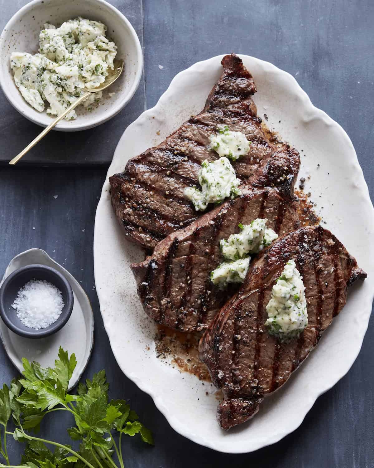 A platter with three grilled rib eye steak topped with a small dollop of compound butter, and a bowl of compound butter and a little bowl of salt on the side.