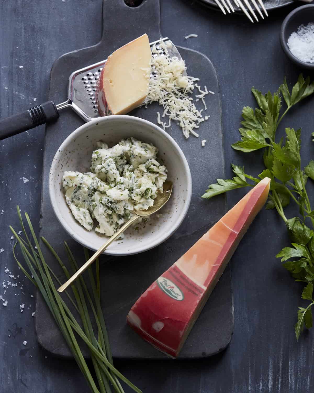 A cheese board with a bowl of compound butter, a grater with a block of cheese partially grated, another block of cheese, some parsley and chives, along with a little bowl of salt.