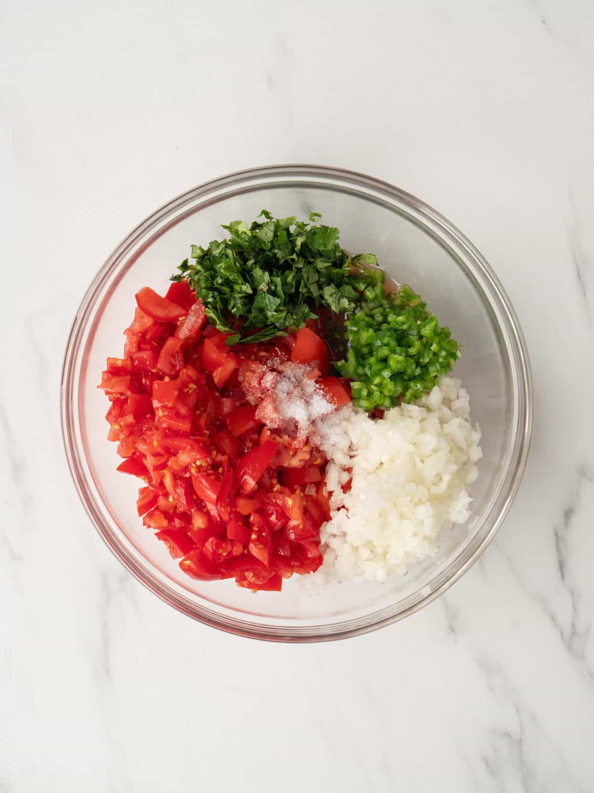 A glass mixing bowl with ingredients to make pico de gallo, diced tomatoes, onions, jalapeño, cilantro and lime juice. 