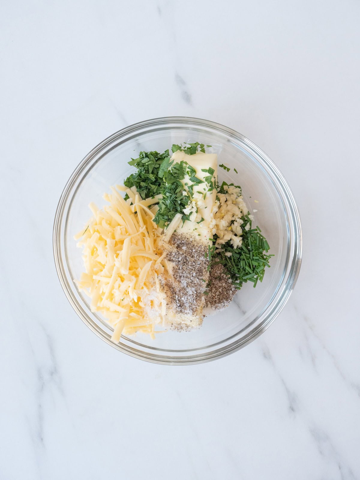A glass mixing bowl with ingredients to make compound butter, which are butter, finely chopped parsley, chopped chives, chopped garlic, shredded cheese, salt and pepper.