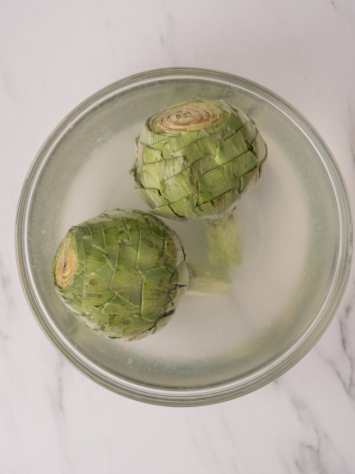 A large glass mixing bowl with a lemon water bath, and two cleaned and prepped artichokes in it.