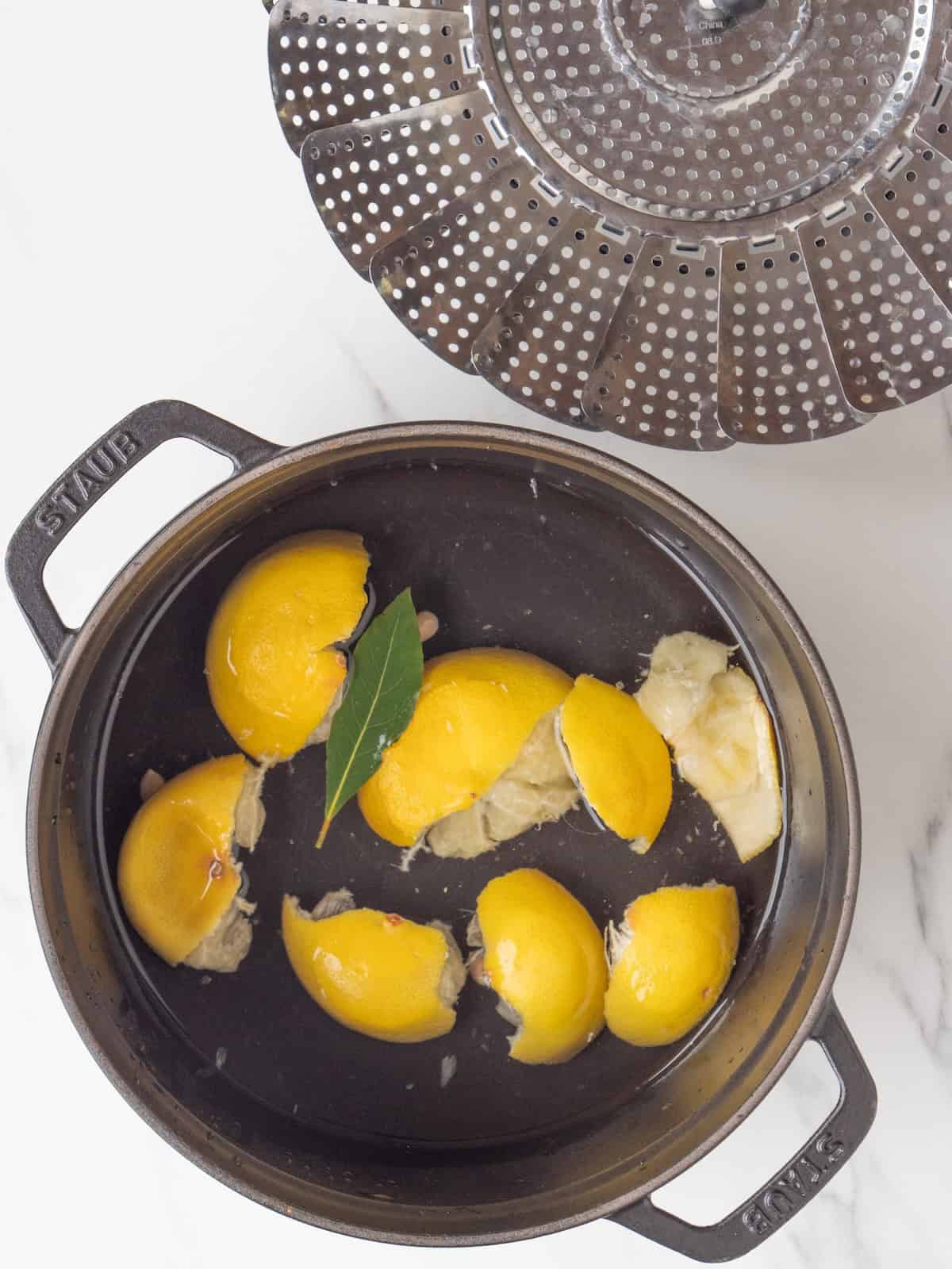 A large pot with about 1-2 inches of water, a bay leaf and lemon peels simmering with a steamer basked on the side.