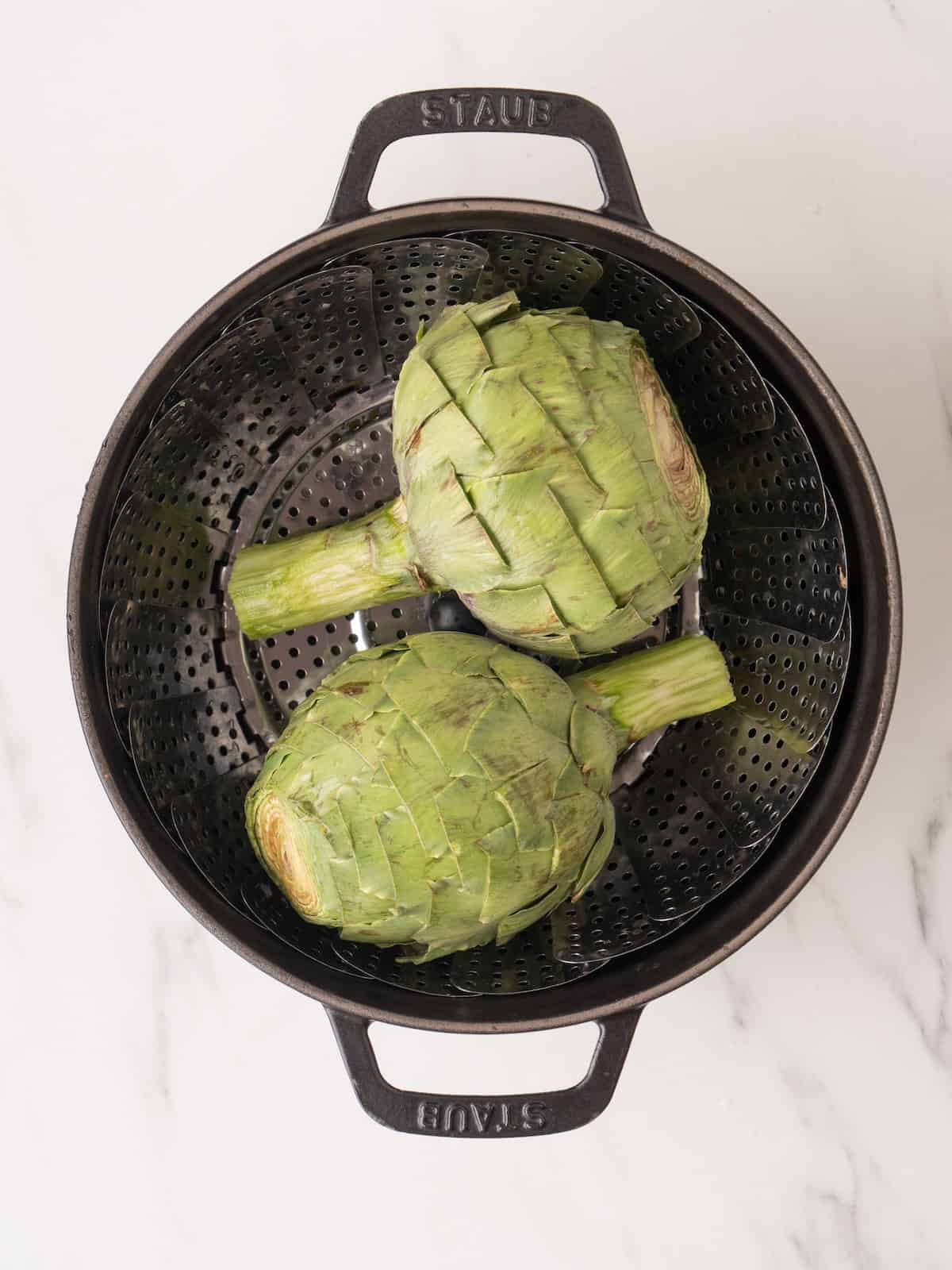 A large pot with a steamer basket in it that has two artichokes being steamed.