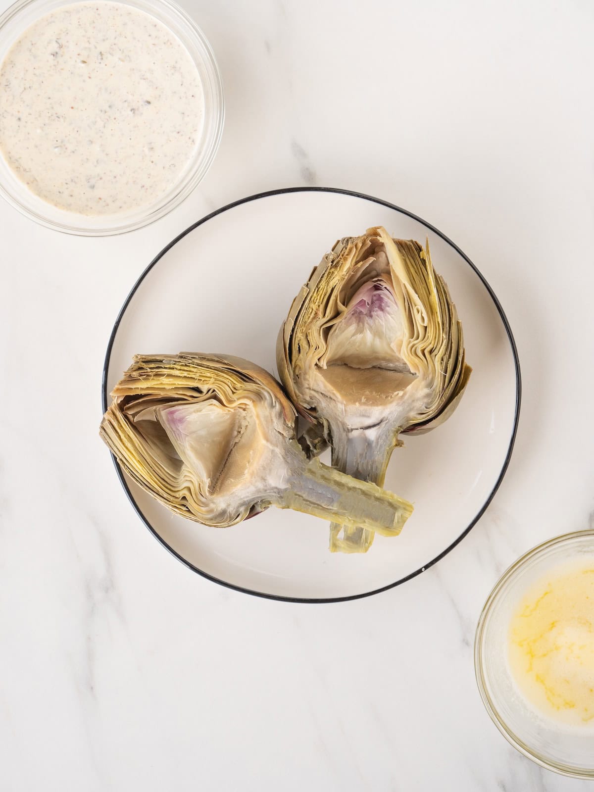 A plate with two halves of a steamed artichoke, along with a small bowl with melted butter and another with caper dipping sauce on the side.