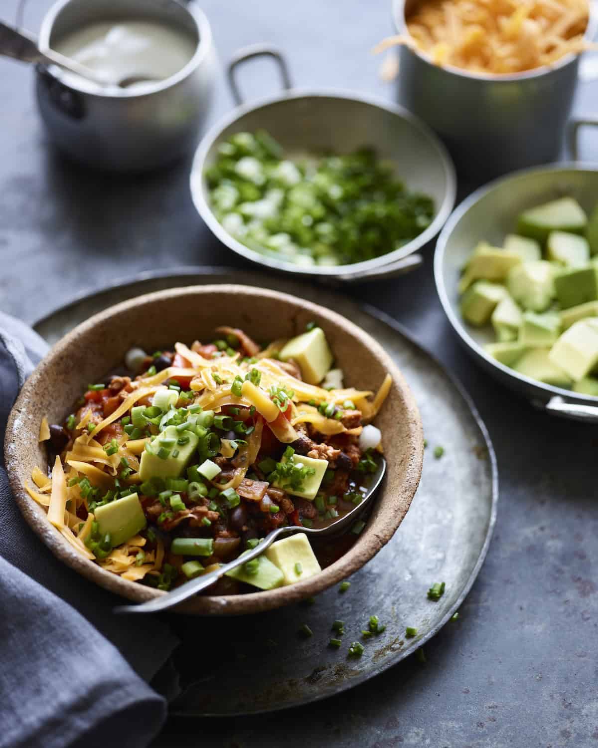 A bowl with chipotle chicken chili topped with avocado, scallions and shredded cheese surrounded with bowls containing these toppings and a bowl of sour cream.