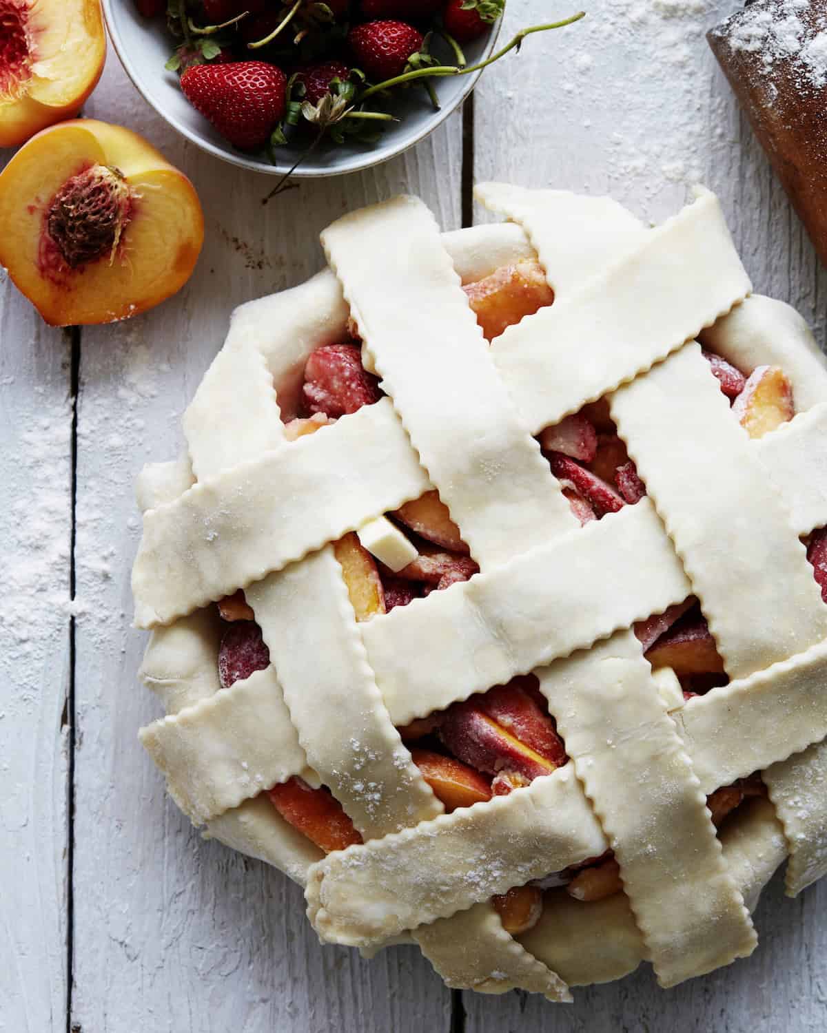 A strawberry peach pie with a lattice top ready to be baked, along with some halved peaches and a bowl of strawberries on the side.