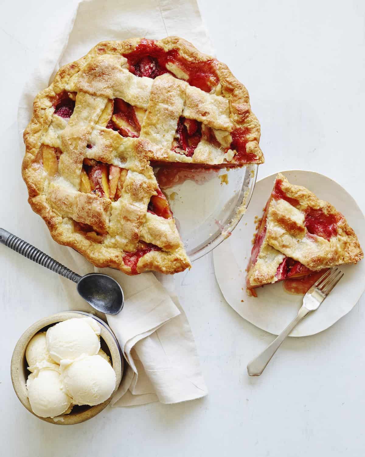 A strawberry peach pie with a lattice top, with a slice cut and placed on a plate on the side, and a bowl with vanilla ice cream scoops on the side.