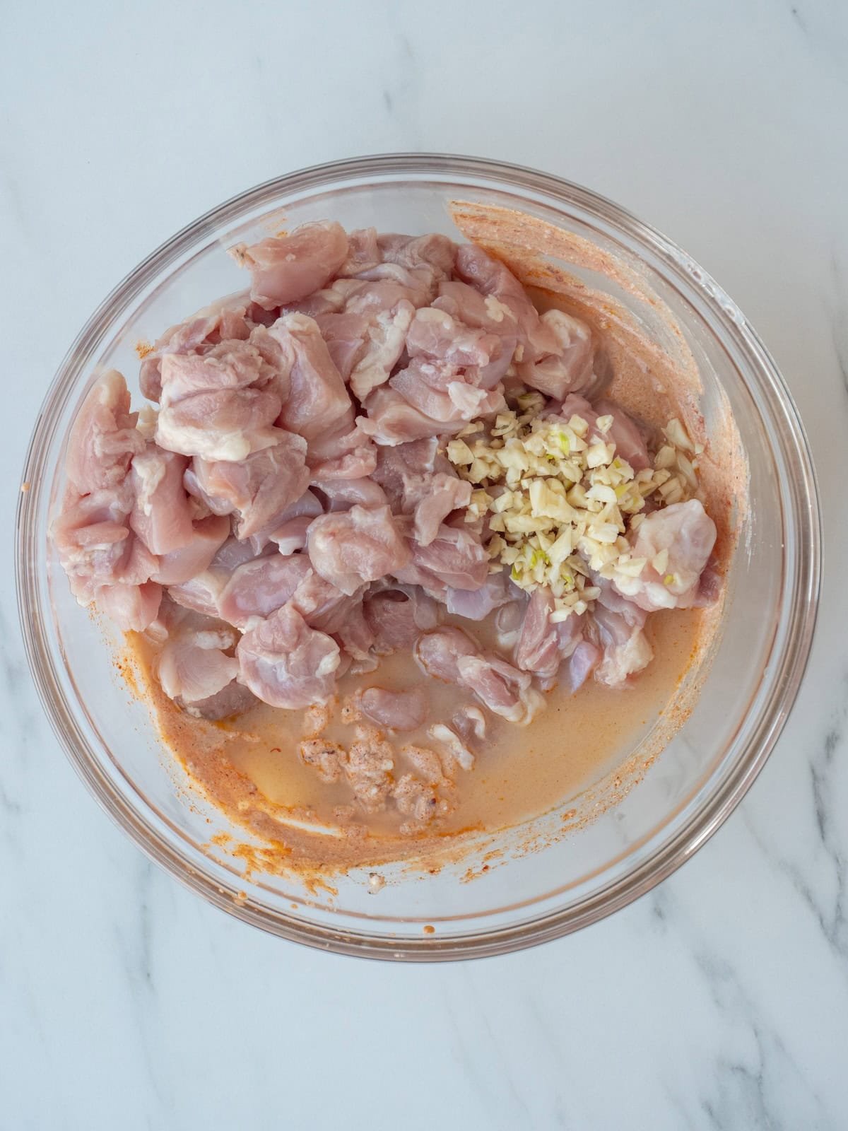 A large glass mixing bowl with chicken cubes added to the yogurt marinade, with garlic and lemon juice added. 
