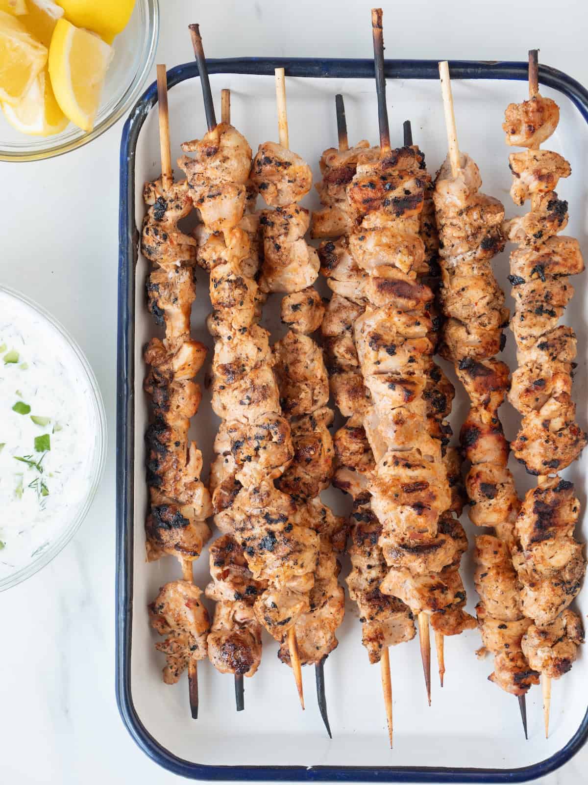 A rectangular dish with grilled chicken skewers with a bowl of tzatziki and a bowl of lemon wedges on the side.