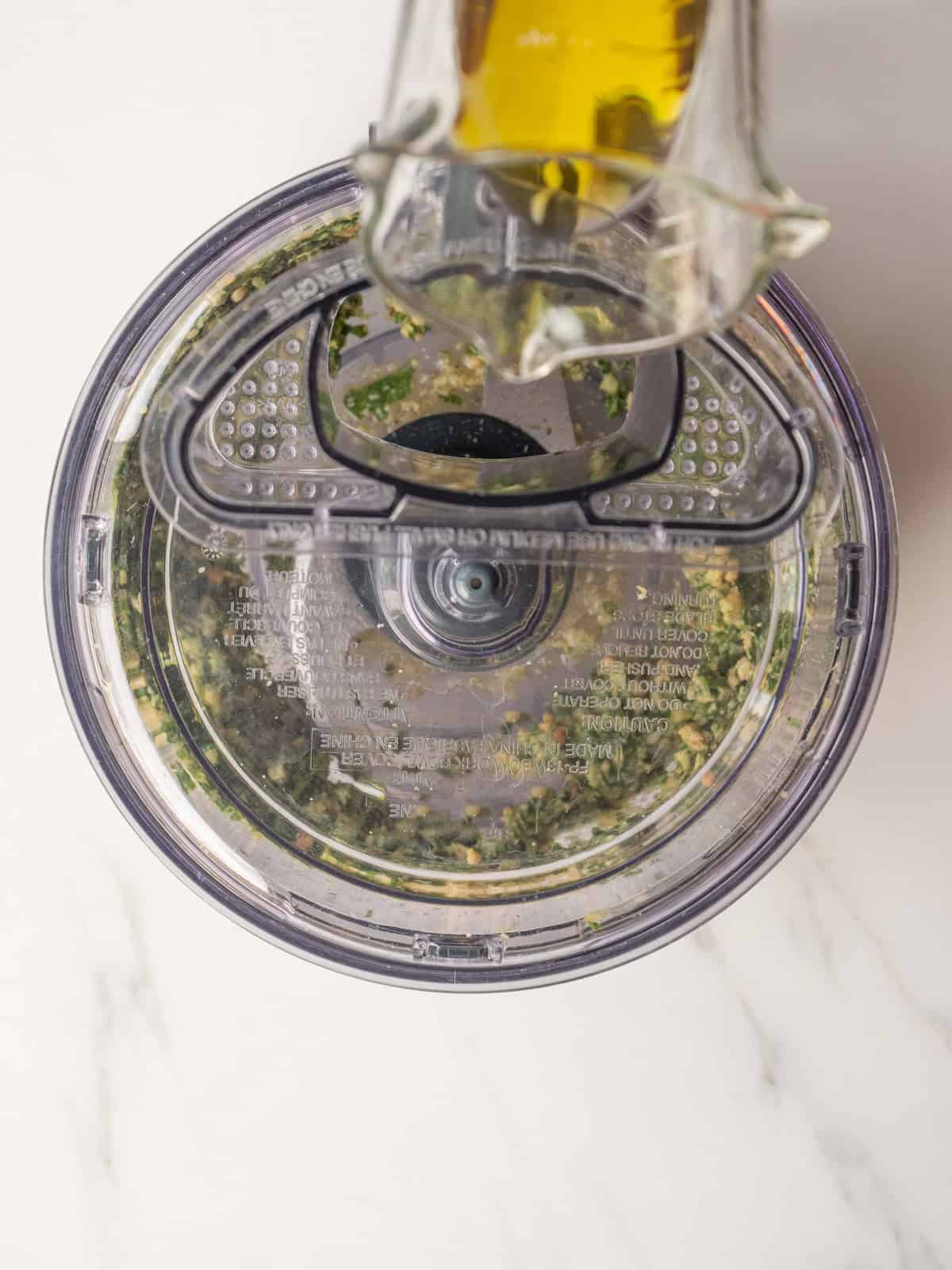 An overhead shot of a food processor with basil pesto being made and olive oil being drizzled into as its blending.