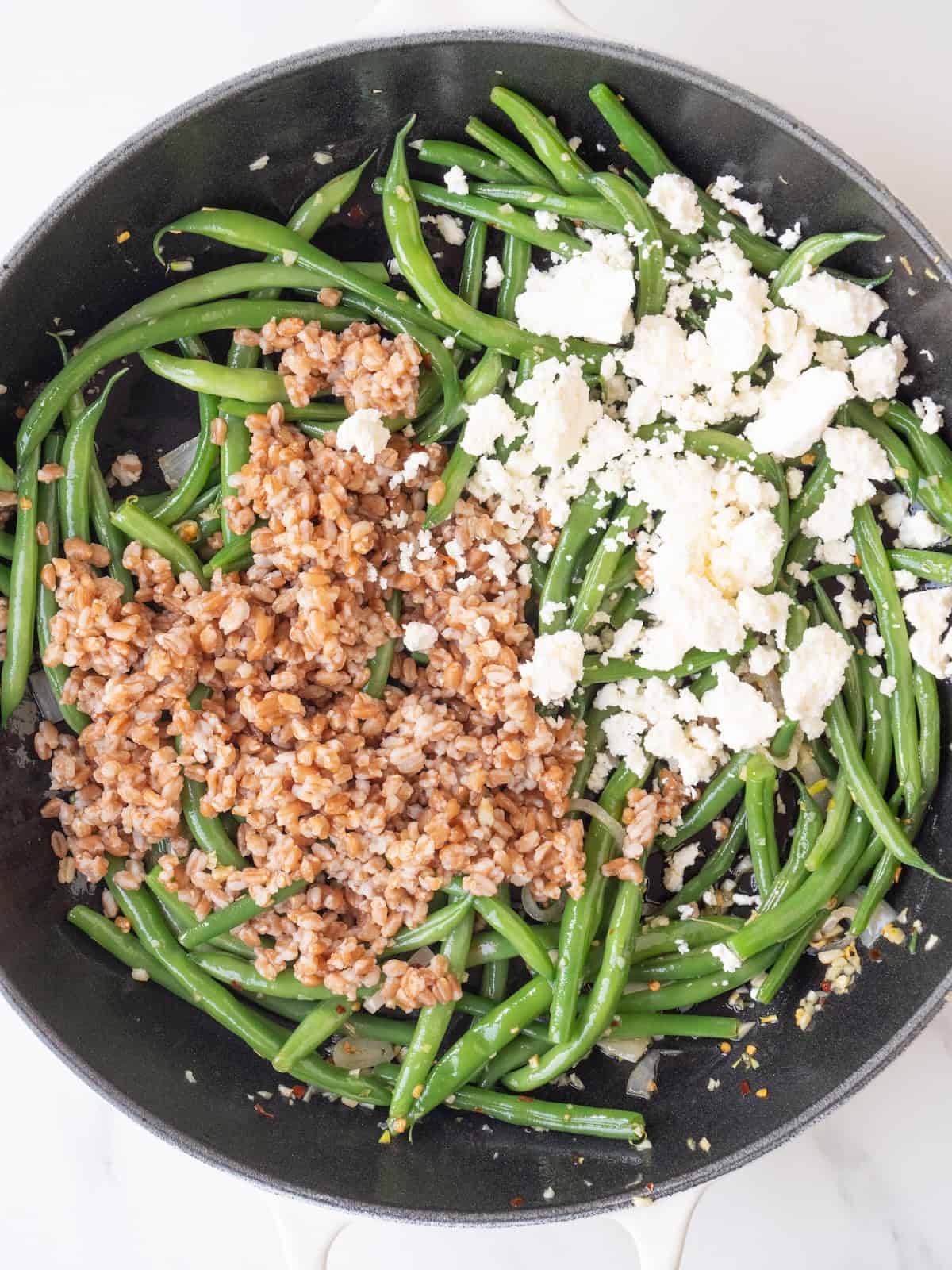 A skillet with green beans being booked, and cooked farro and crumbled feta just added. 
