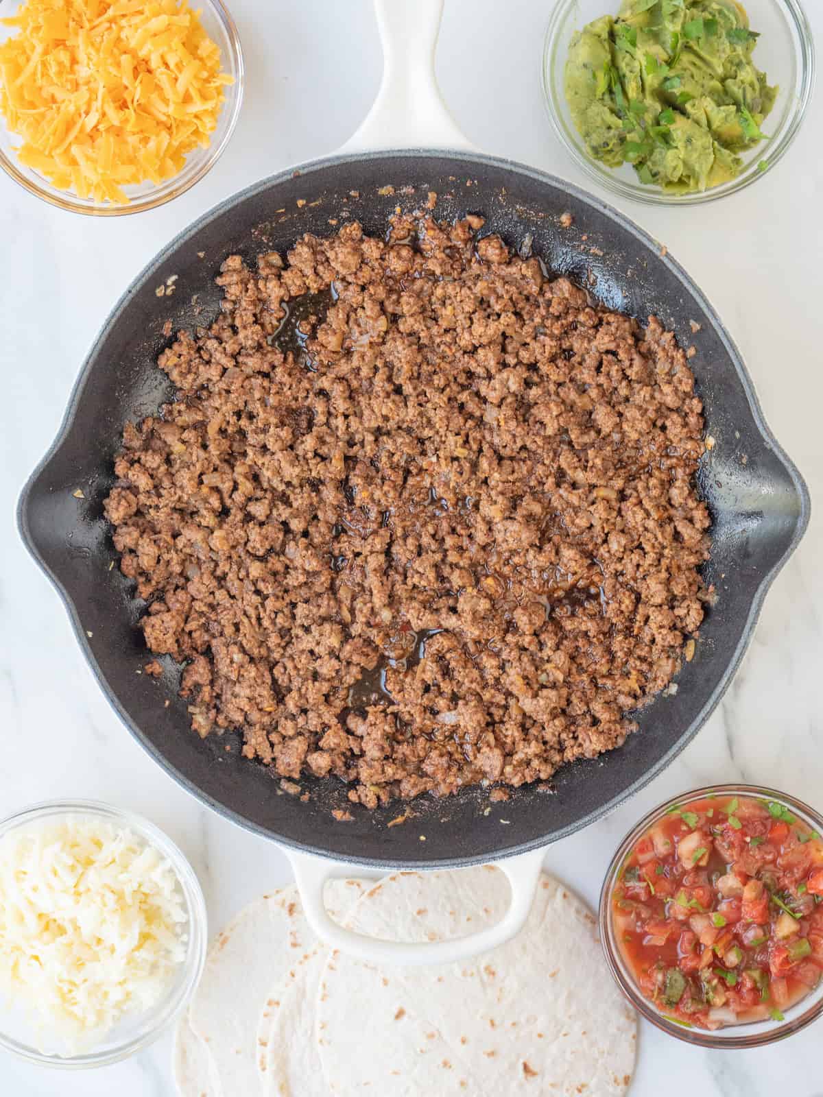 A skillet with ground beef cooked for tacos, along with four small glass mixing bowls with shredded Cheddar cheese, shredded Monterey Jack cheese, guacamole and salsa, along with tortillas on the side.