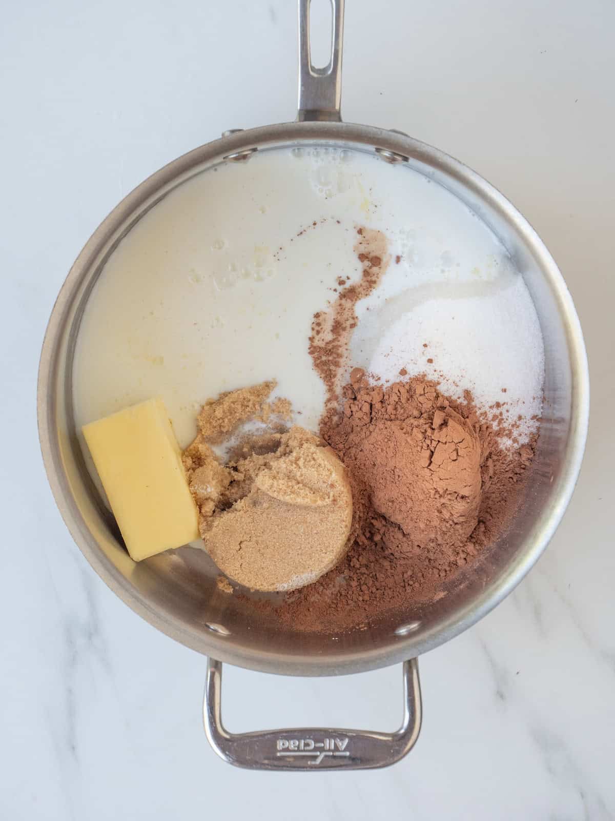 A skillet with white sugar, light brown sugar, butter, cocoa powder being mixed together and melted to form a mix.