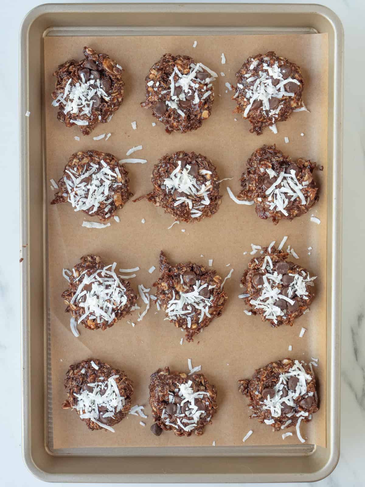 A parchment paper lined baking sheet with mounds of the no bake cookies, topped with chocolate chips and coconut flakes and flattened slightly.