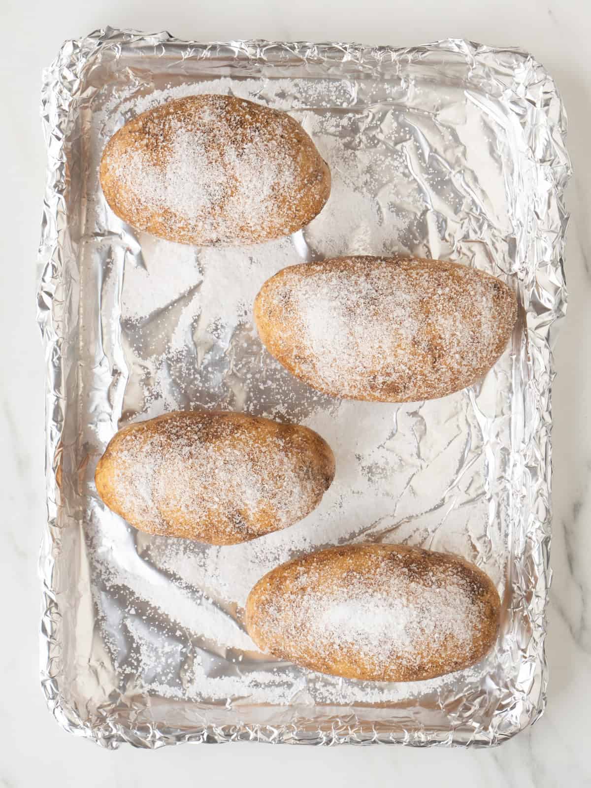 A baking sheet pan lined with foil with four russet potatoes rubbed with salt ready to be baked.