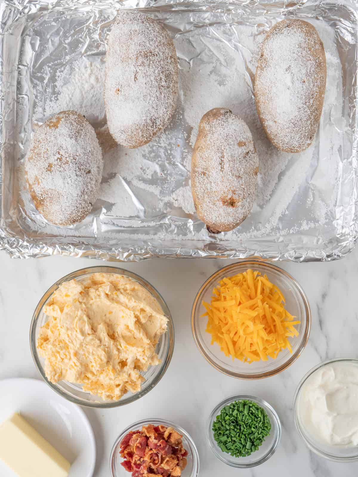 A baking sheet pan lined with foil with four baked potatoes rubbed with salt, along with several bowls of toppings on the side- garlic cheese butter, shredded cheese, chopped chives, sour cream, bacon bits and a stick of butter.