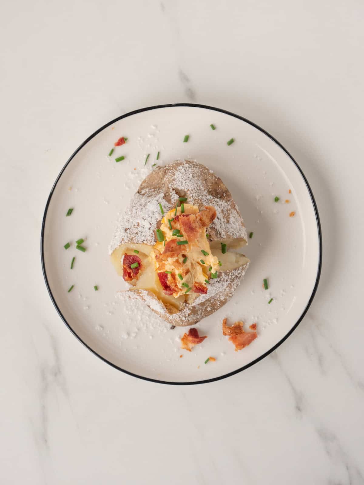 A plate with a baked potato rubbed with salt and topped with garlic cheese butter, bacon bits and chives.