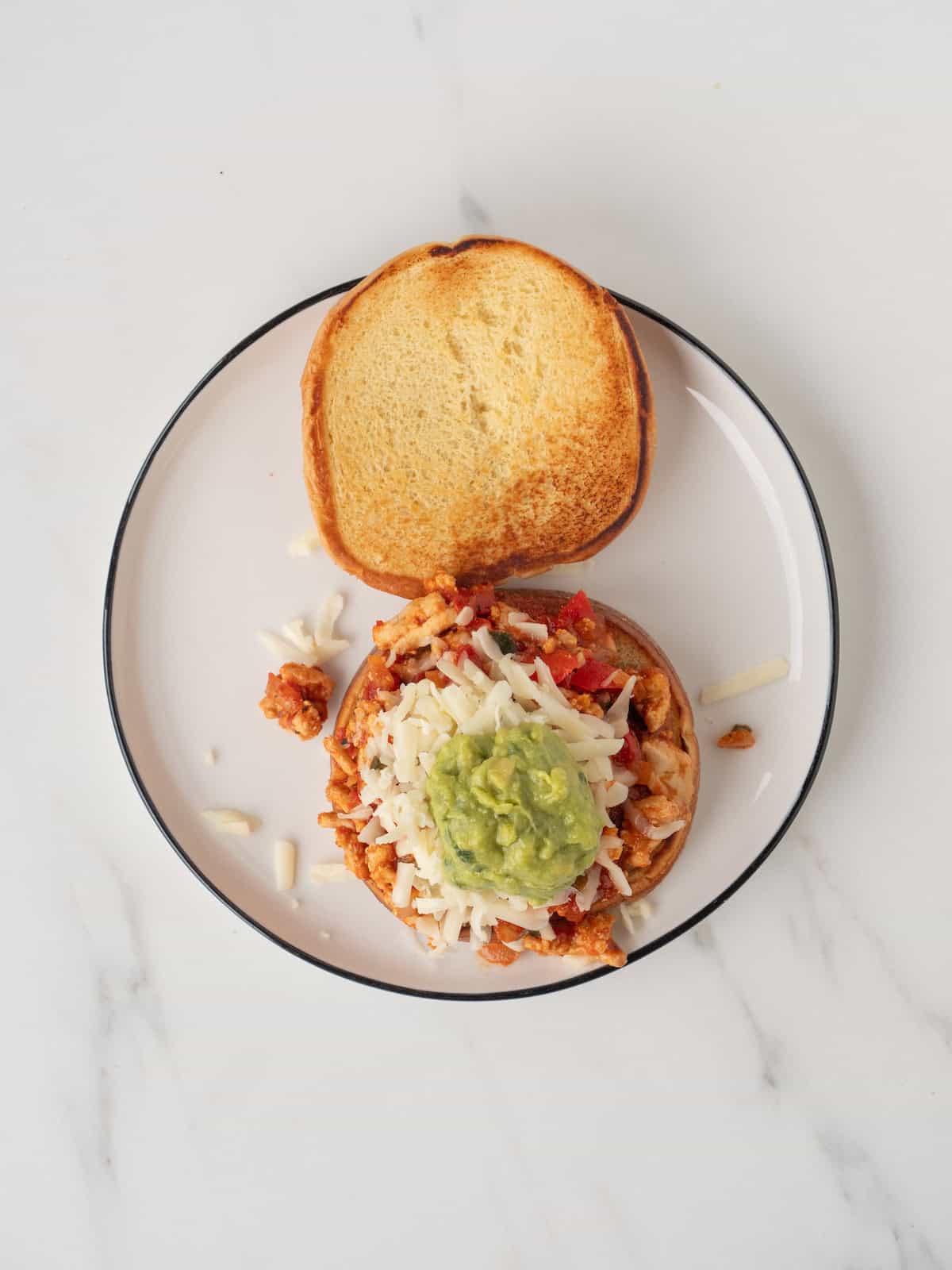 A plate with a tex mex style sloppy joe assembled on a bun, with a ground turkey filling topped with shredded cheese and guacamole, with the top of the bun on the side.